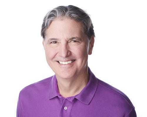 A smiling person with gray hair wearing a purple polo shirt against a white background.