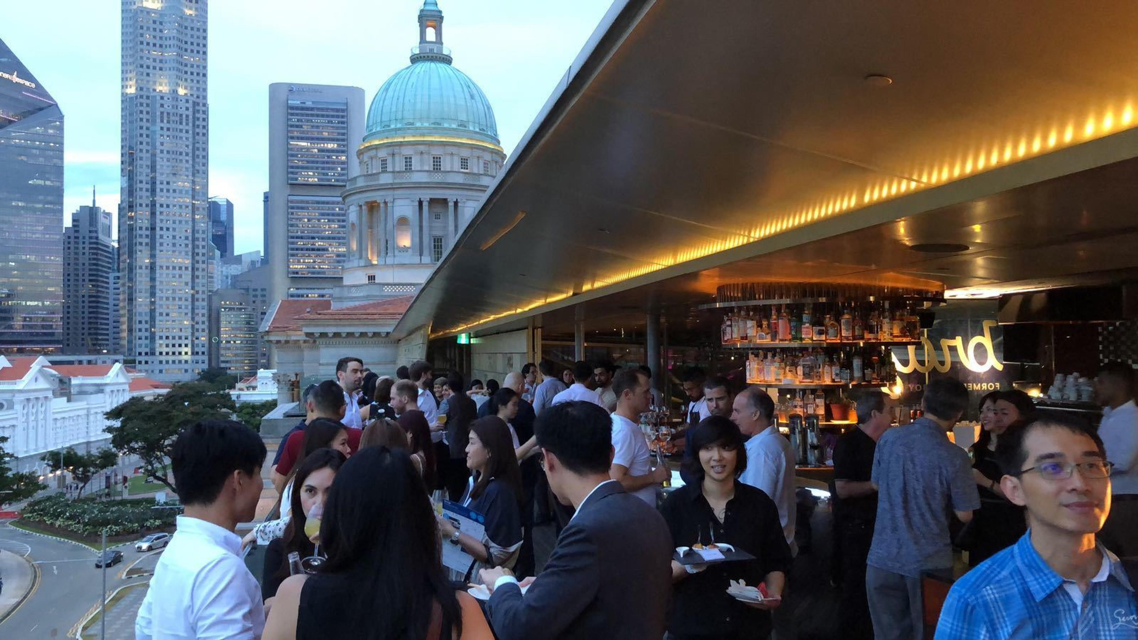 A bustling rooftop bar with people socializing and enjoying drinks, set against a backdrop of city skyscrapers and a historic dome building at dusk.