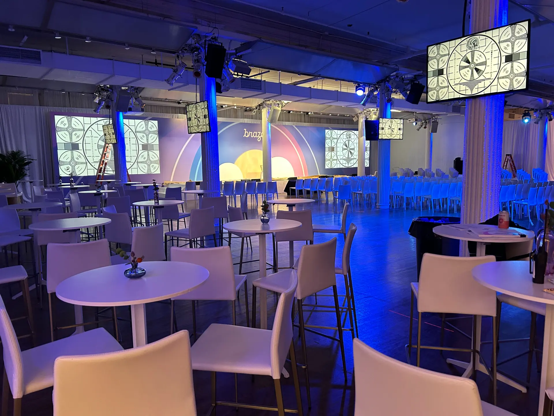 Sala de eventos con mesas y sillas blancas, decorada con luces azules. Pantallas en las paredes muestran gráficos y un escenario al fondo.