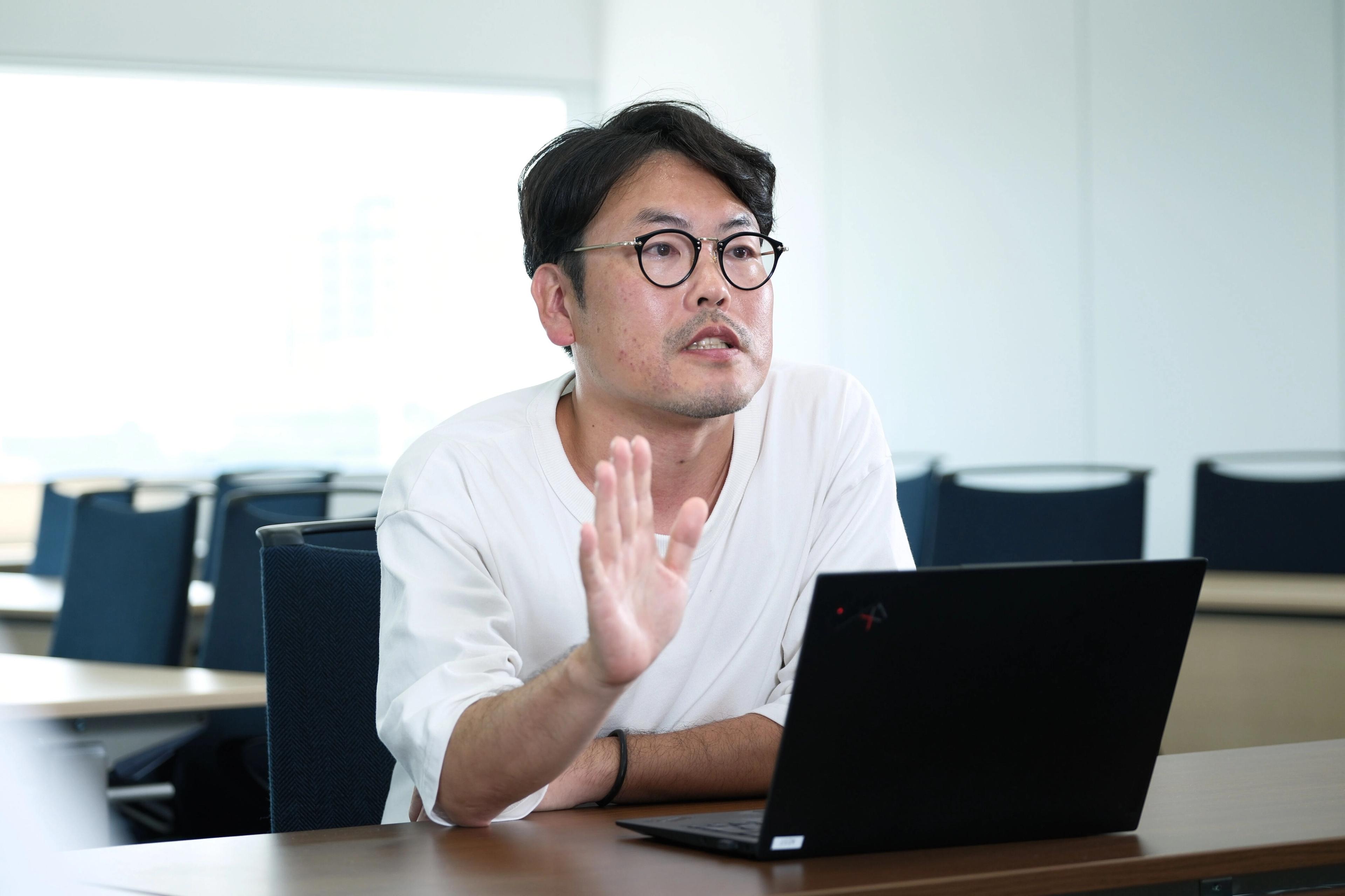 男性がノートパソコンの前で話している様子。手を挙げて説明をしています。