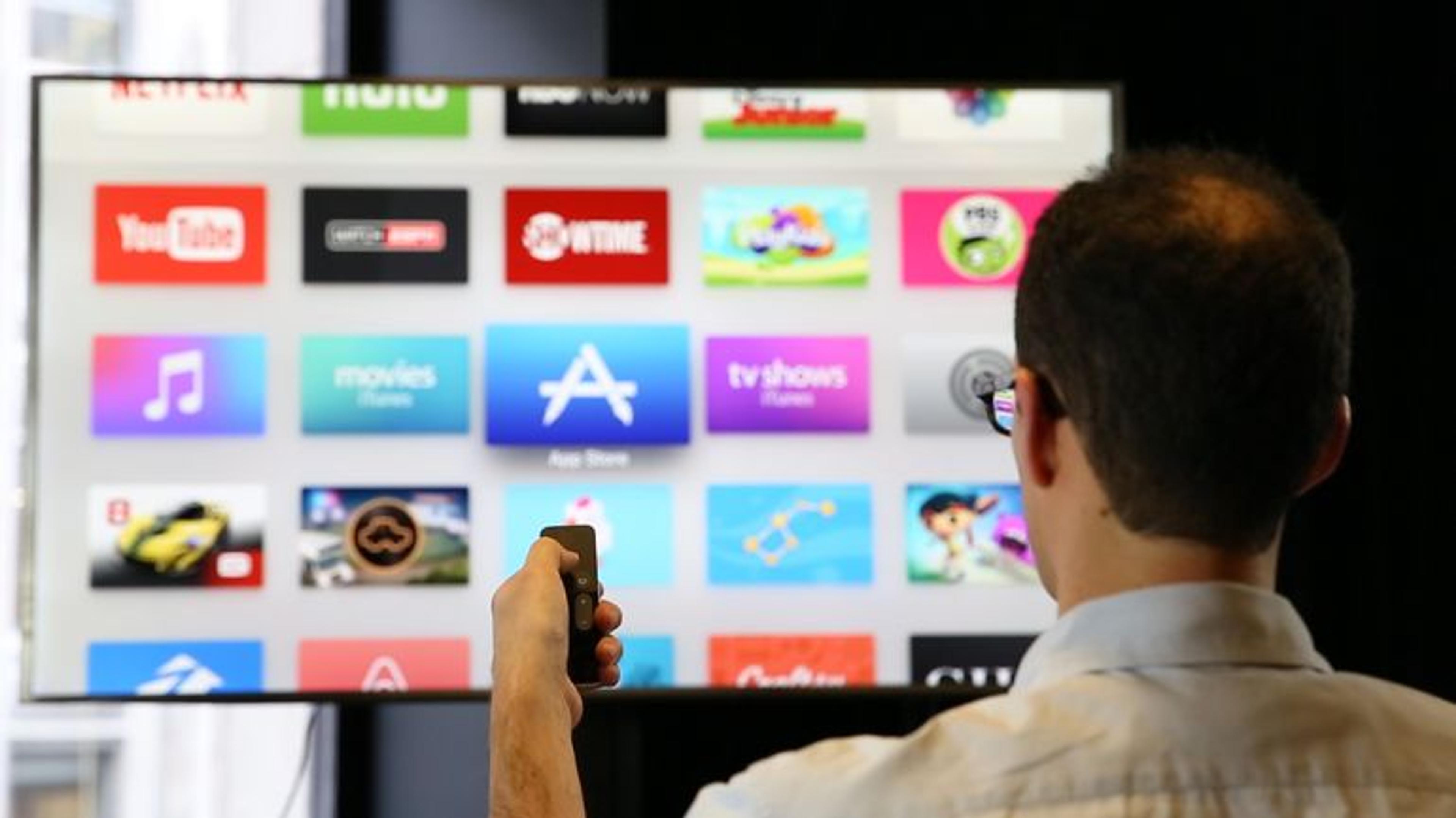 A person holding a remote control faces a television displaying various app icons, including YouTube and Netflix. The setting appears to be indoors with a dark background.