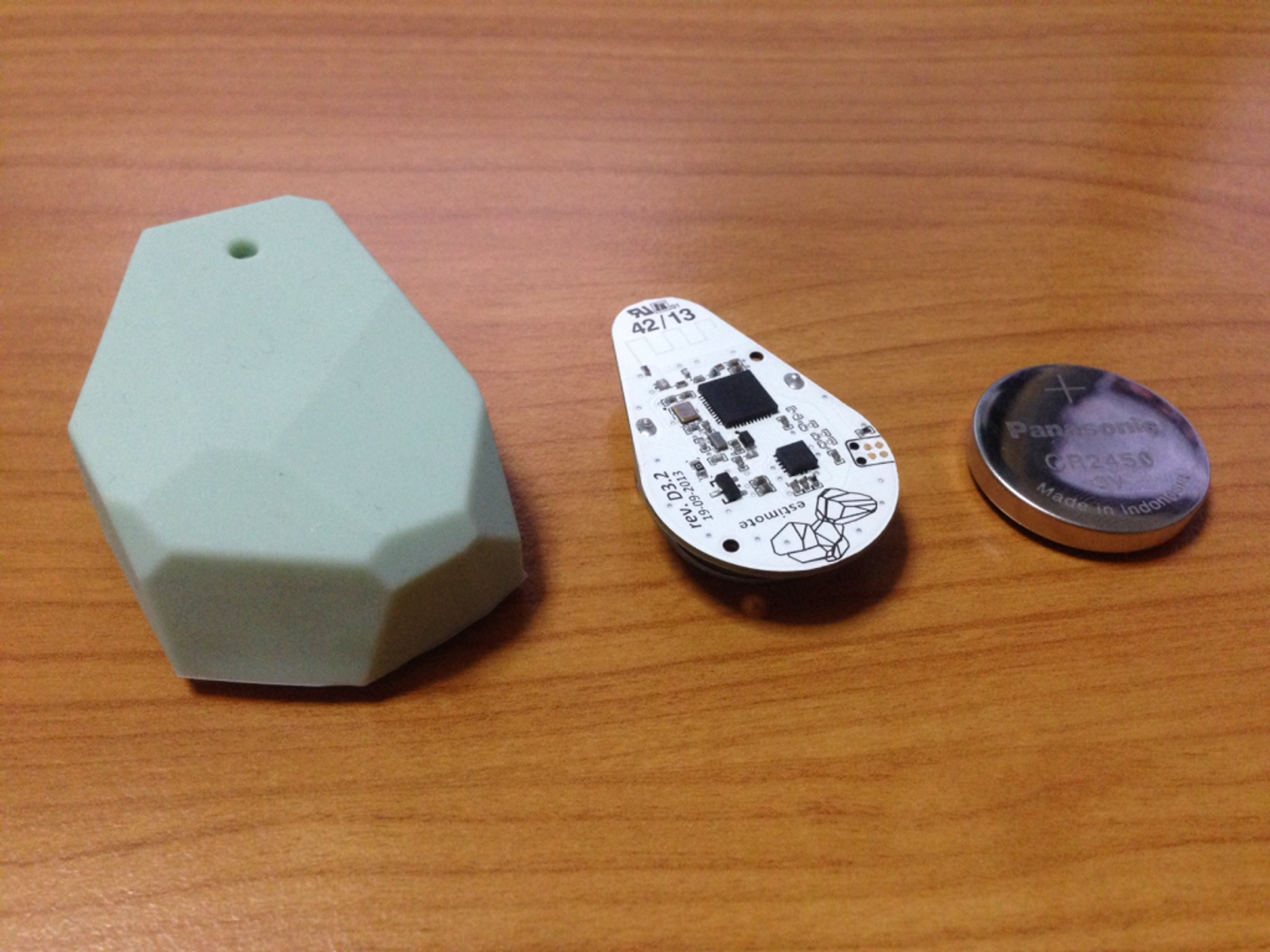 A light green silicone casing, a circuit board, and a round battery are displayed on a wooden surface. The items are arranged from left to right.