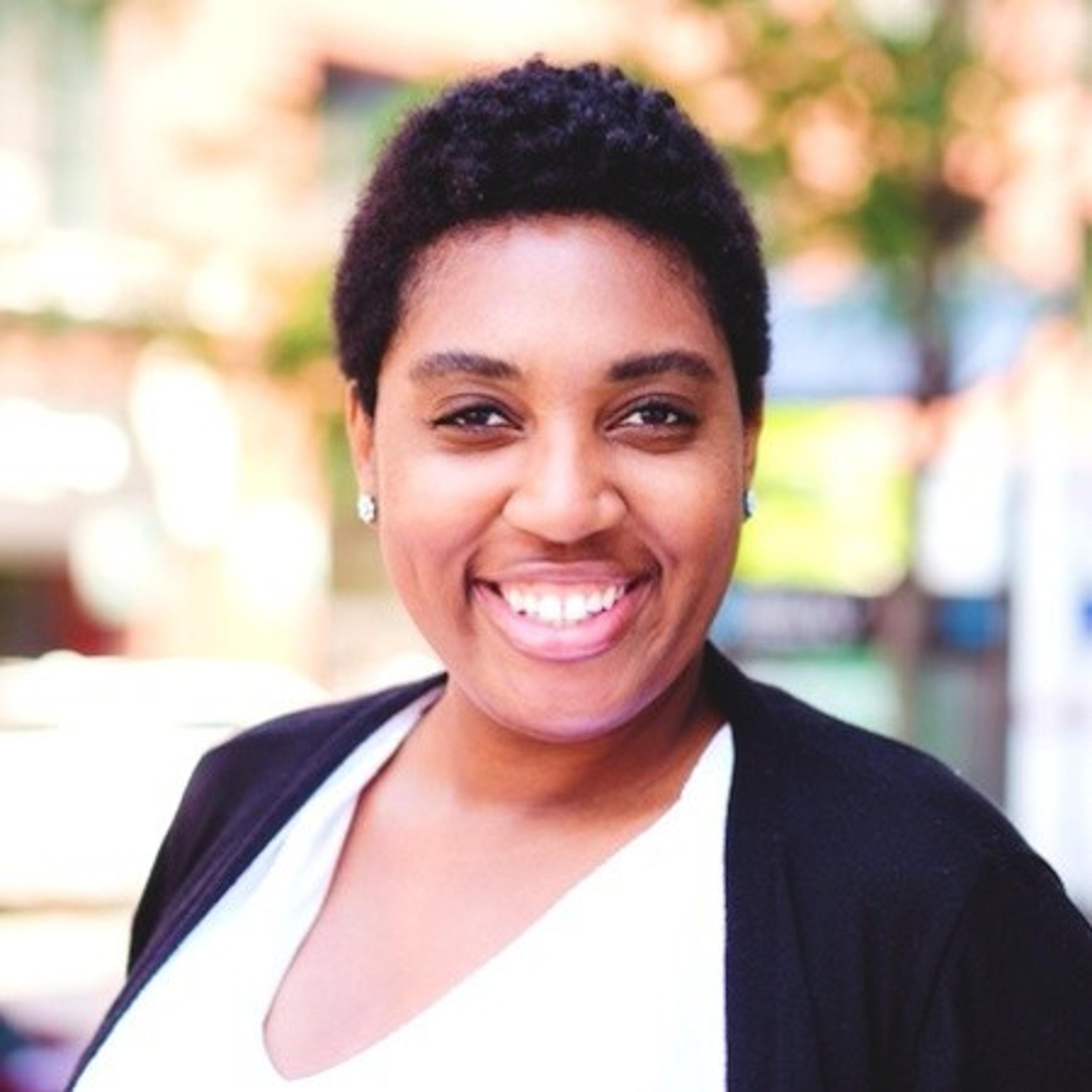 A smiling woman with short, curly hair stands outdoors, wearing a black cardigan over a white shirt. Soft sunlight highlights her cheerful expression.