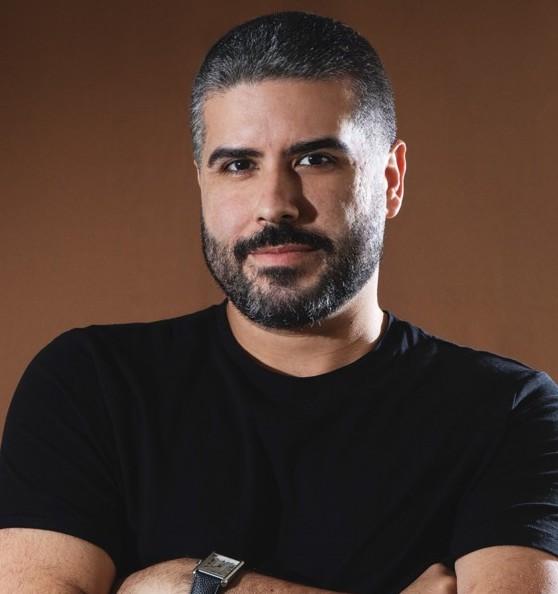 A man with short gray hair and a beard is smiling confidently while wearing a black shirt. He has his arms crossed and is set against a brown background.