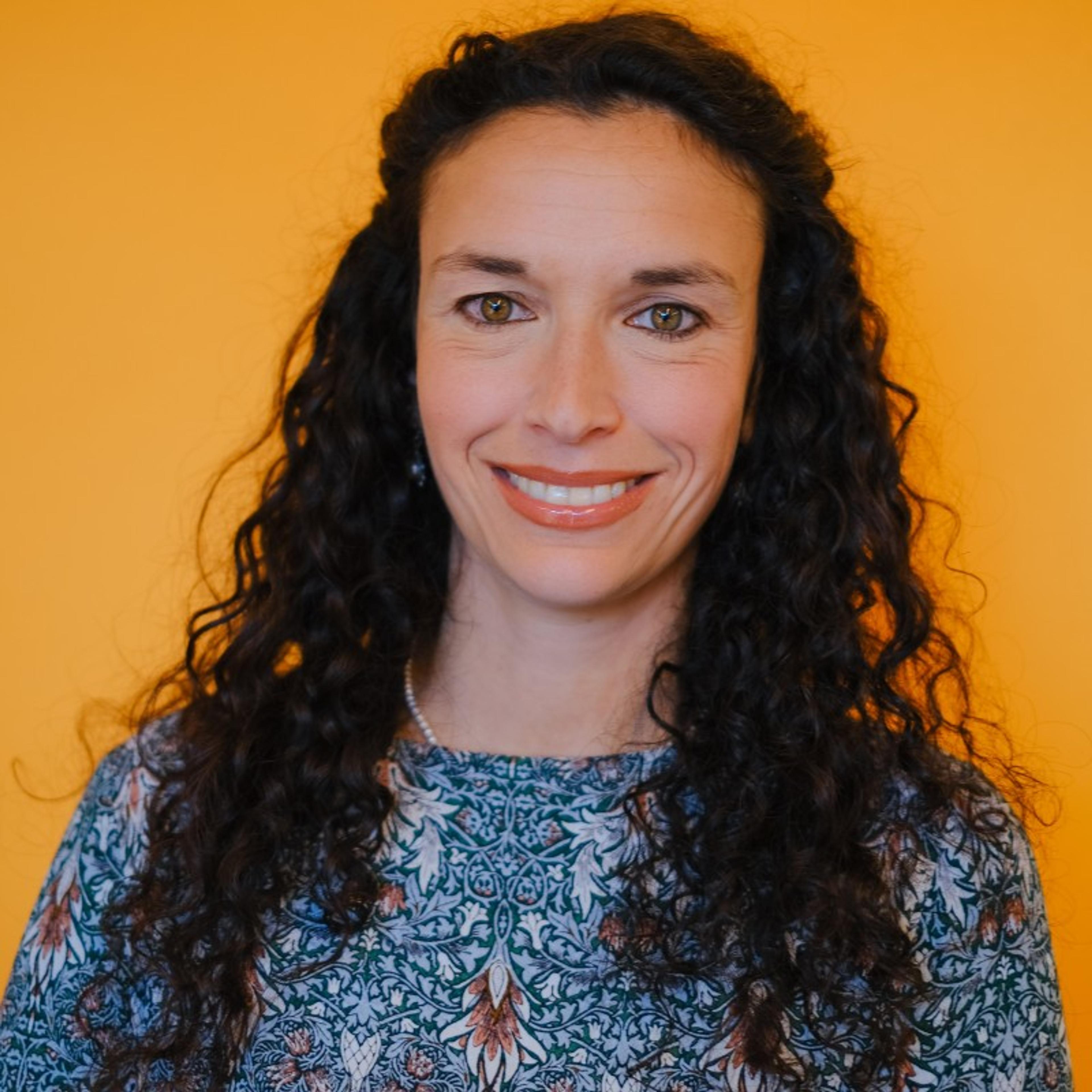 Headshot of a smiling woman with long, dark curly hair against a warm orange background.