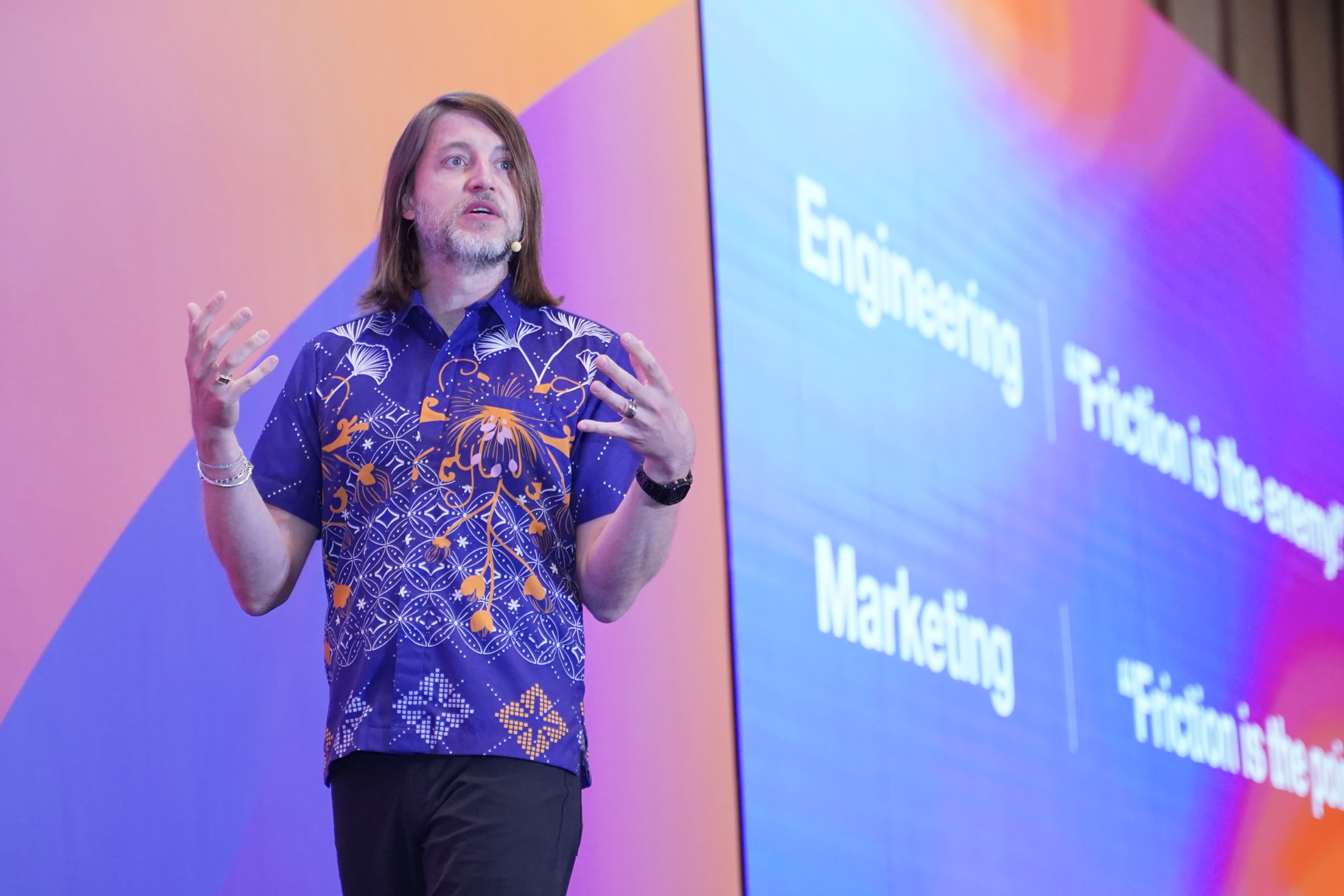 A speaker wearing a colorful patterned shirt gestures onstage beside a presentation screen at a conference.