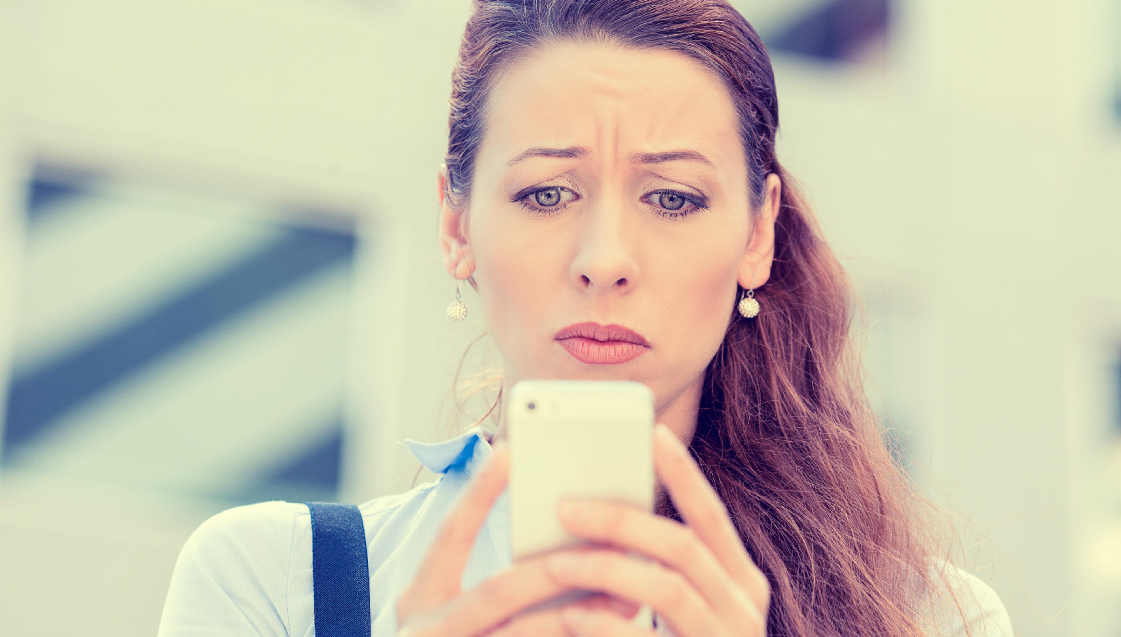 A woman with long brown hair looks worried while checking her smartphone. She appears concerned and is wearing a light blue shirt and earrings.