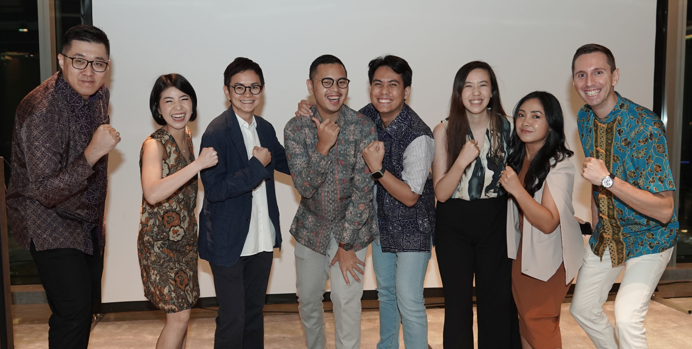A group of eight people stands together, smiling and posing with their fists raised in a celebratory manner. They are dressed in a mix of formal and traditional attire.