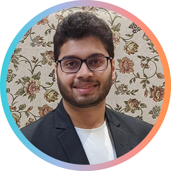 A young man with glasses and a beard smiles, wearing a black blazer over a white shirt. The background features a floral pattern.