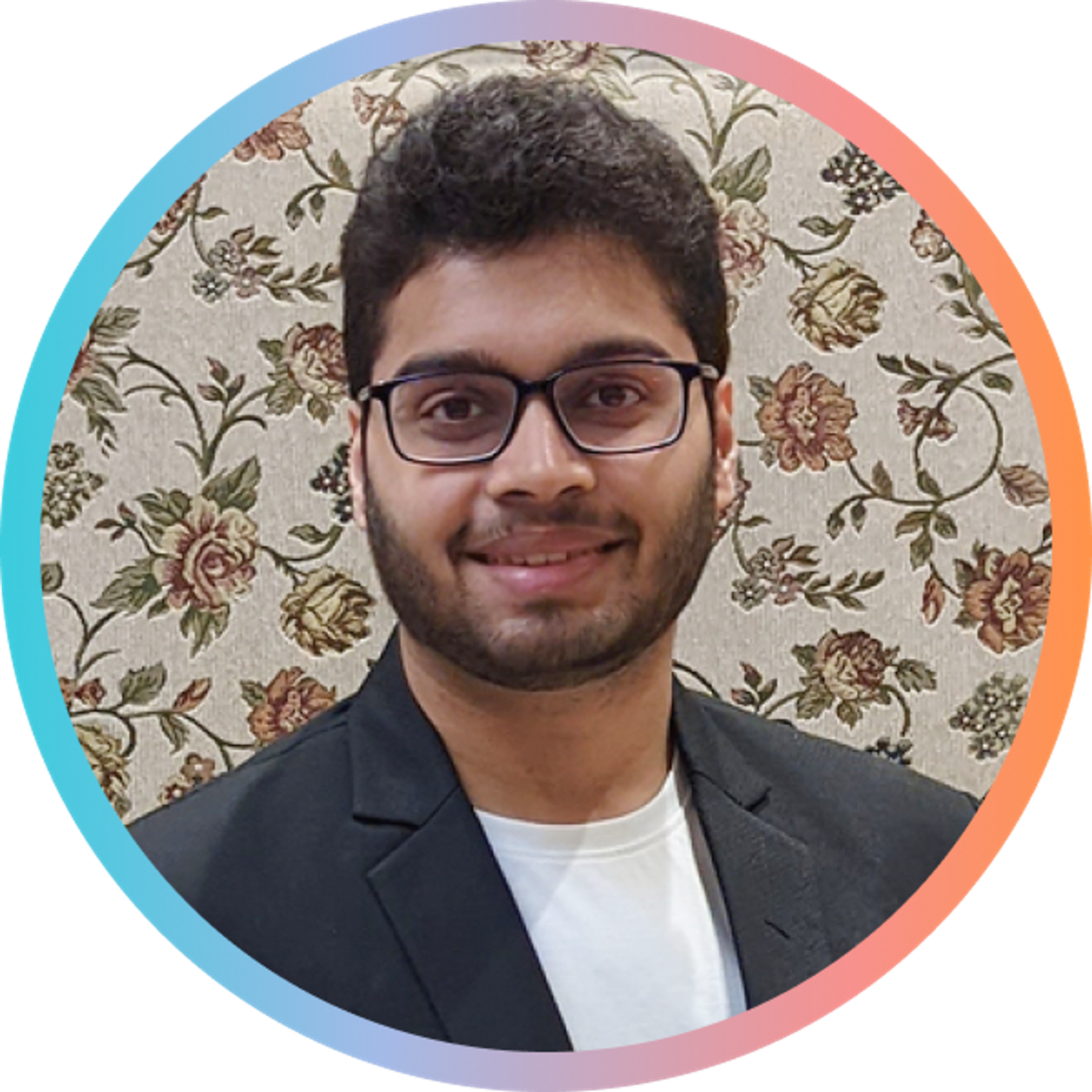 A young man with glasses and a beard smiles, wearing a black blazer over a white shirt. The background features a floral pattern.