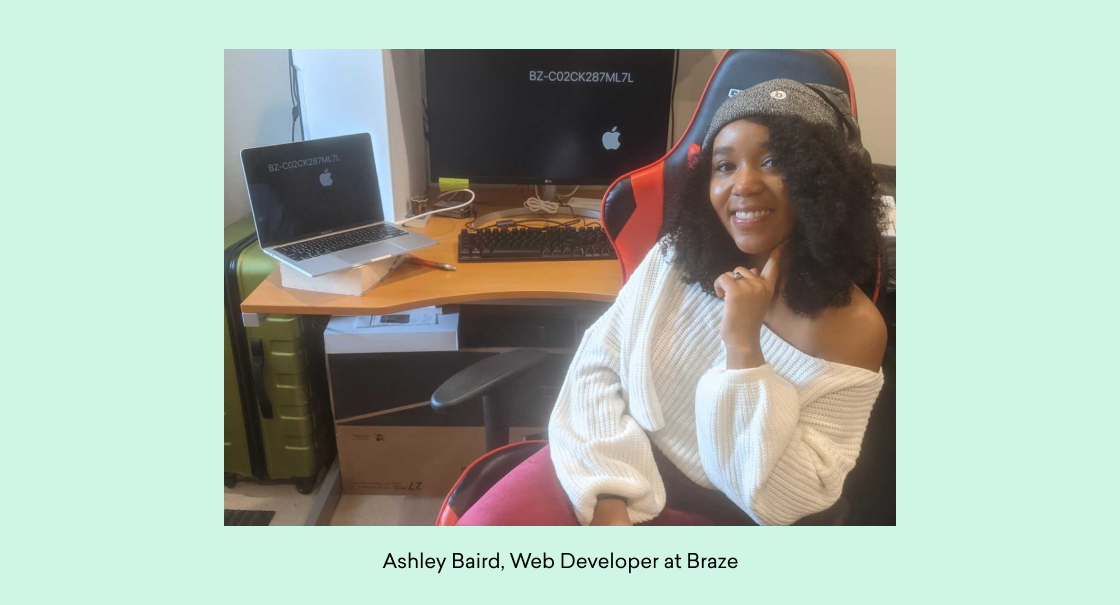 A woman with curly hair sits in an office chair, smiling at the camera. She is wearing a white sweater and is in front of a desk with a laptop and computer monitor.