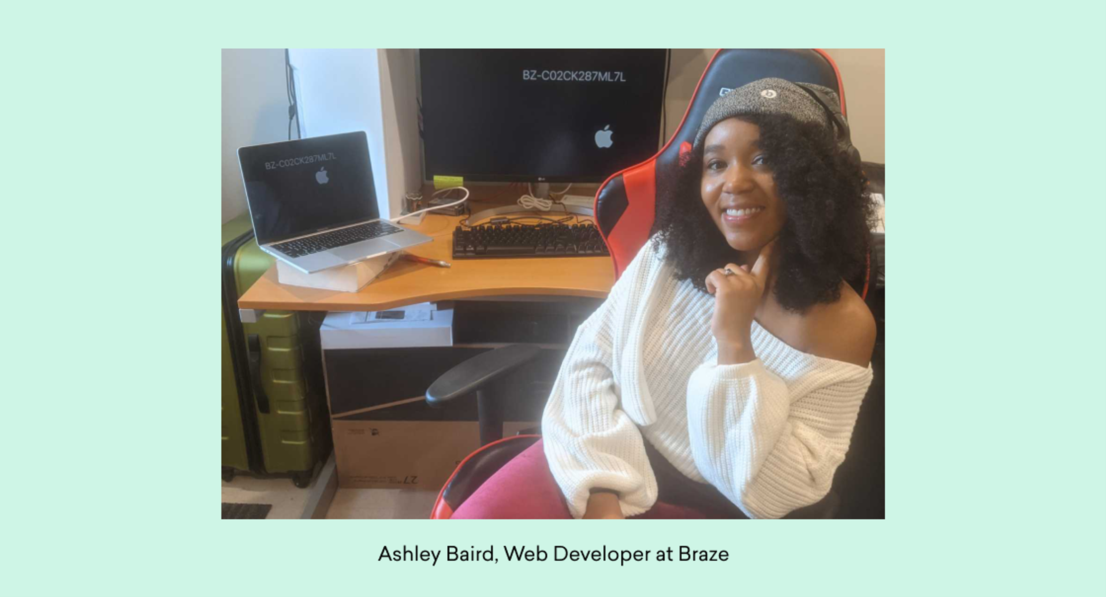 A woman with curly hair sits in an office chair, smiling at the camera. She is wearing a white sweater and is in front of a desk with a laptop and computer monitor.