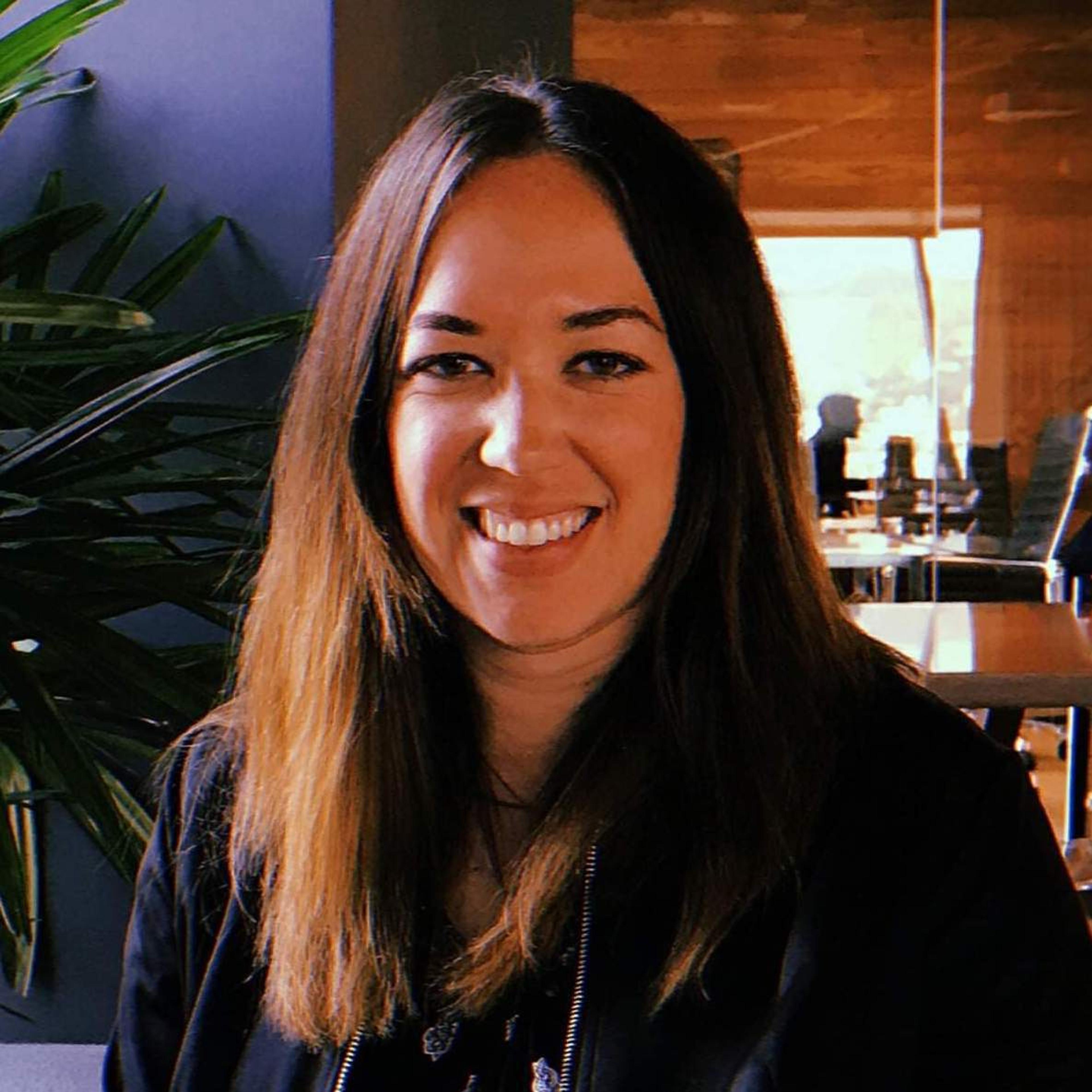 A woman with long, dark hair smiles at the camera, sitting in a modern indoor space with plants in the background.