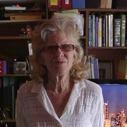 An older woman with glasses stands in front of a bookshelf filled with books and various items. She is wearing a white button-up shirt and smiling at the camera.