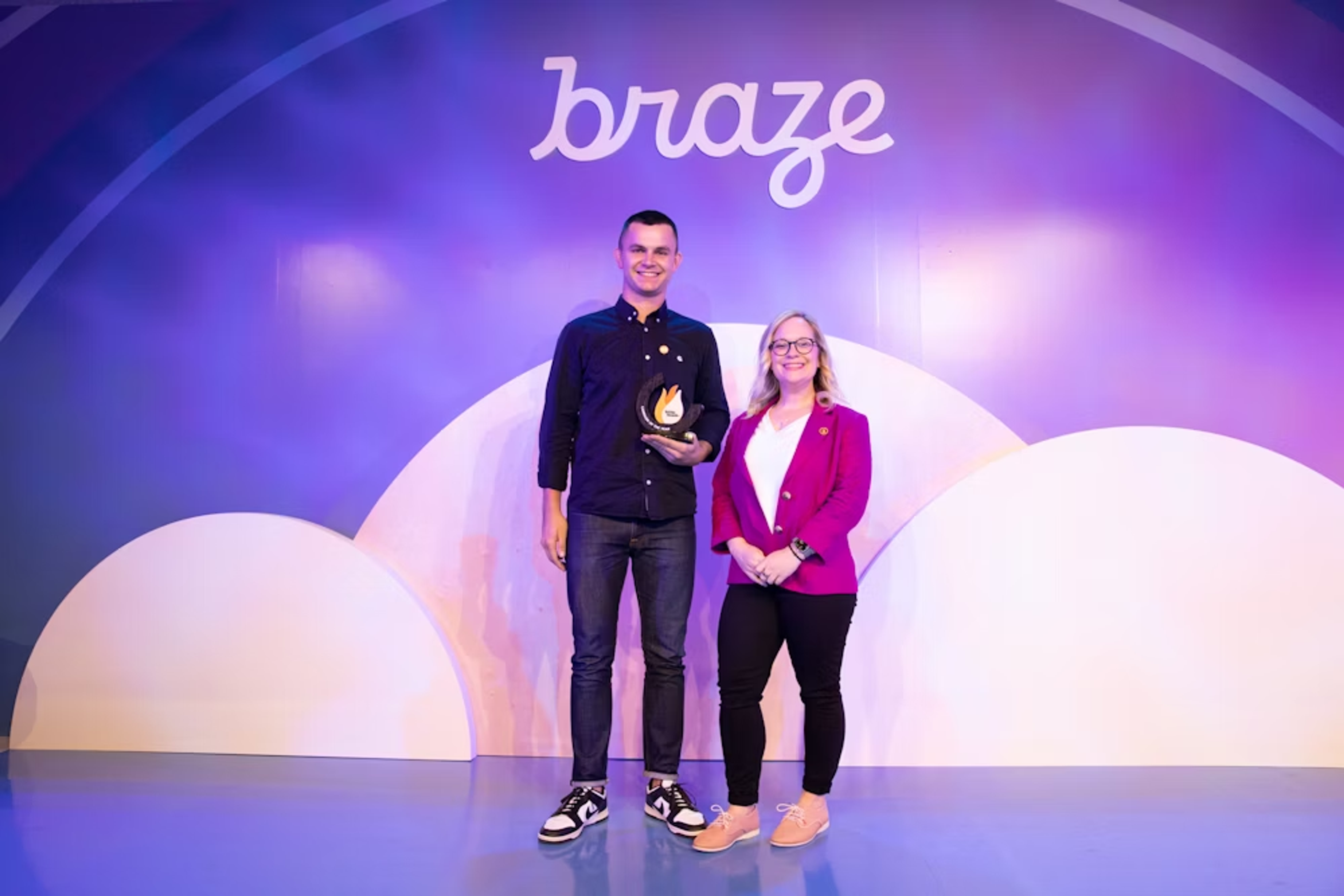 Dos personas de pie frente a un fondo colorido con el logo de "braze". Una persona sostiene un trofeo y la otra sonríe a su lado.