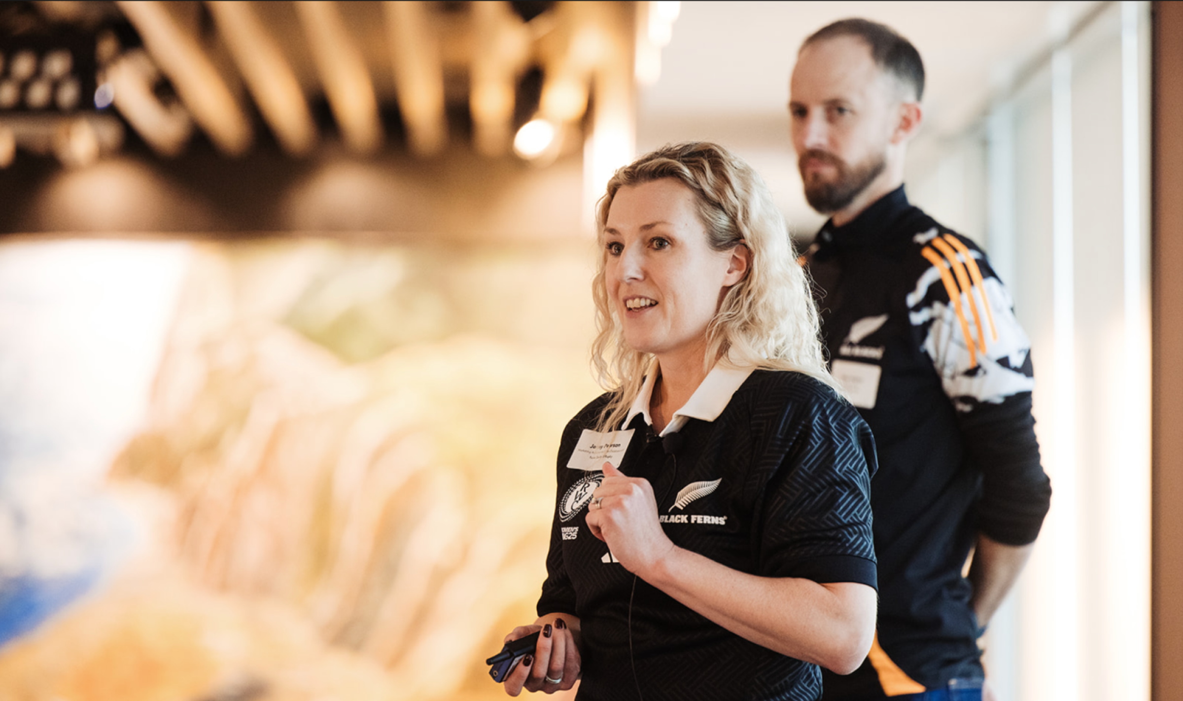 A blonde woman in a black Black Ferns polo shirt speaks, with a bearded man in an Adidas shirt standing behind her.