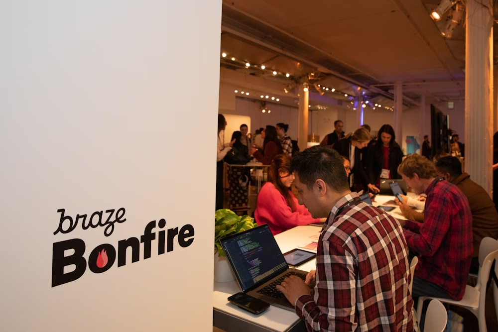 Un grupo de personas trabajando en computadoras portátiles en un evento, con un letrero que dice "braze Bonfire" en primer plano. Hay plantas y una atmósfera de colaboración.