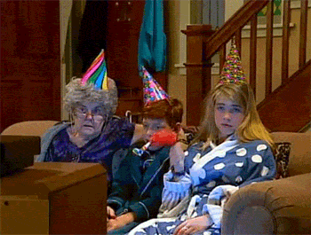 A woman with gray hair and a party hat sits next to a boy in a party hat, while a girl in a polka dot robe holds a red object. They all look surprised while watching television.