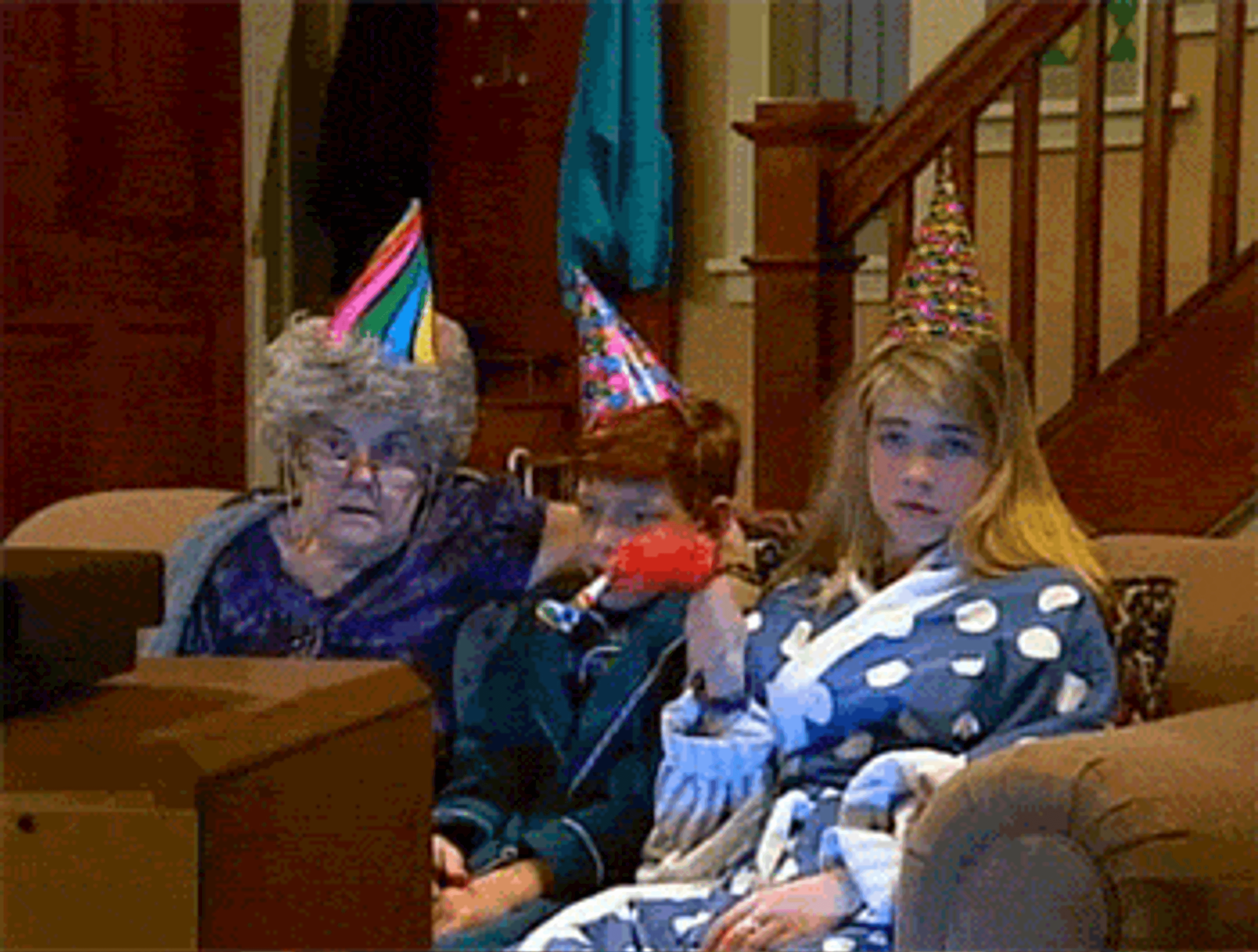 A woman with gray hair and a party hat sits next to a boy in a party hat, while a girl in a polka dot robe holds a red object. They all look surprised while watching television.