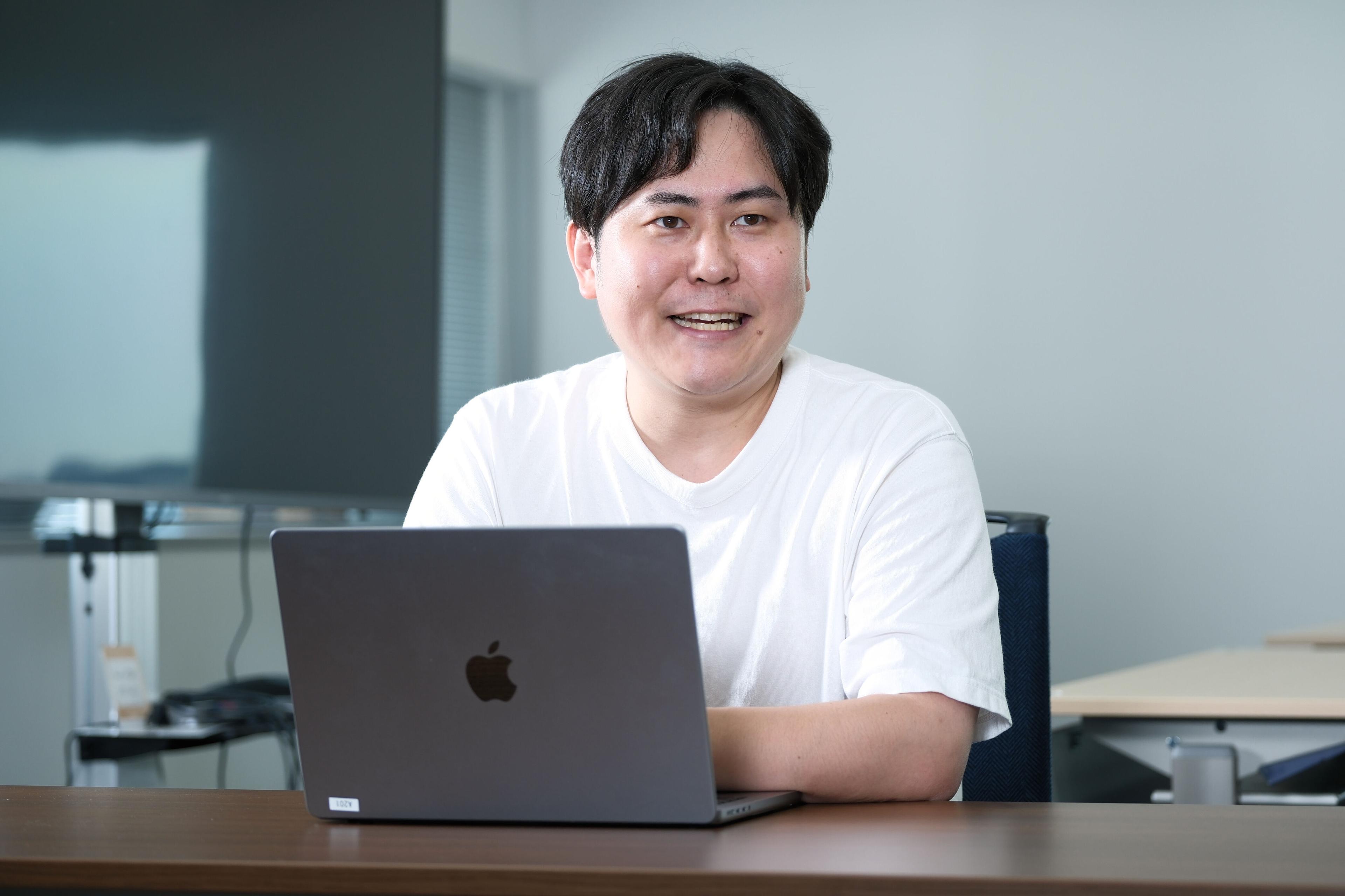 男性がノートパソコンの前で微笑んでいる。背景にはオフィスの一部が見える。