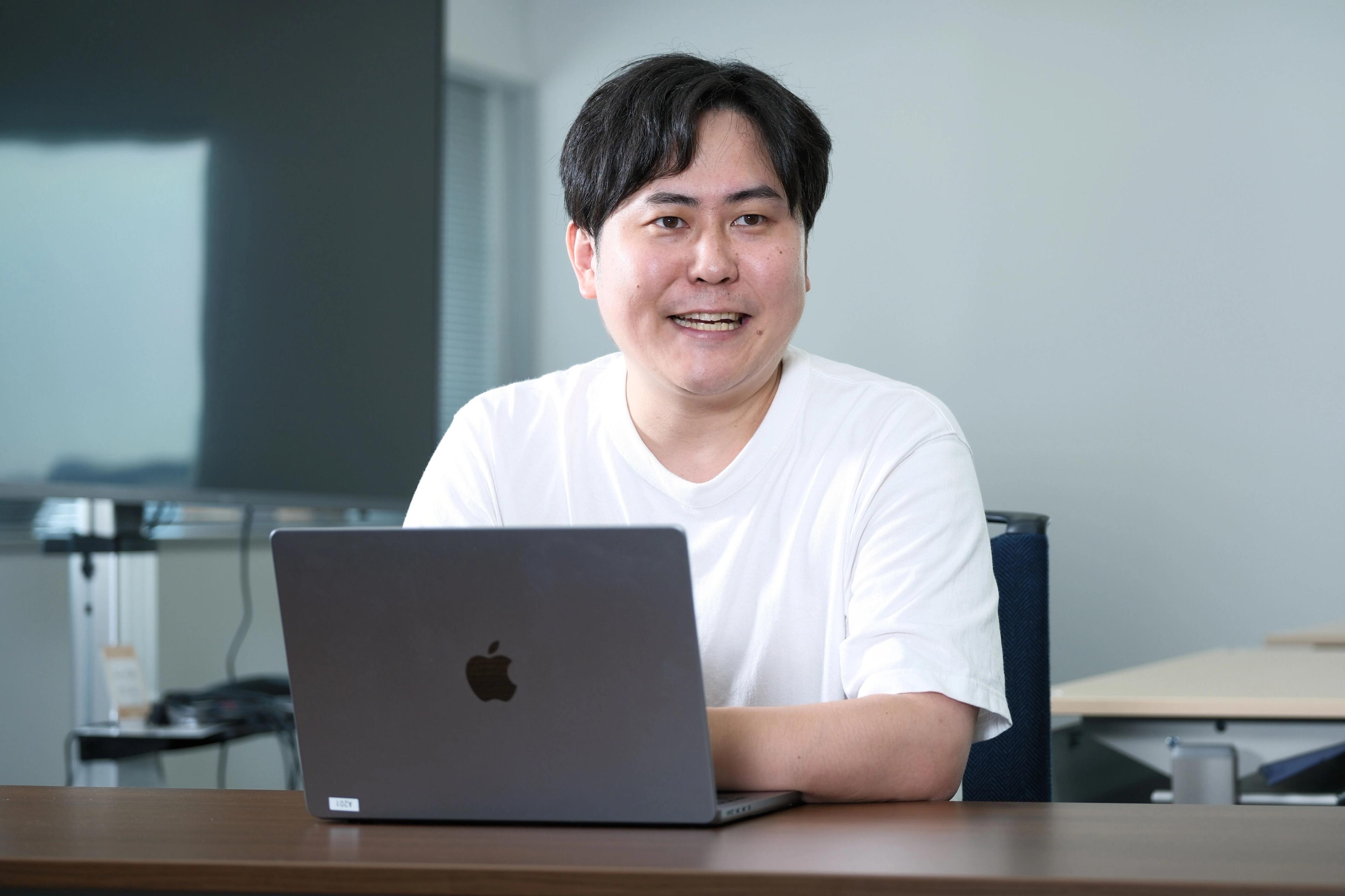 男性がノートパソコンの前で微笑んでいる。背景にはオフィスの一部が見える。