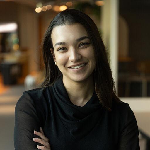 A young woman with long dark hair smiles confidently while crossing her arms. She is wearing a black top and is in a well-lit indoor setting.
