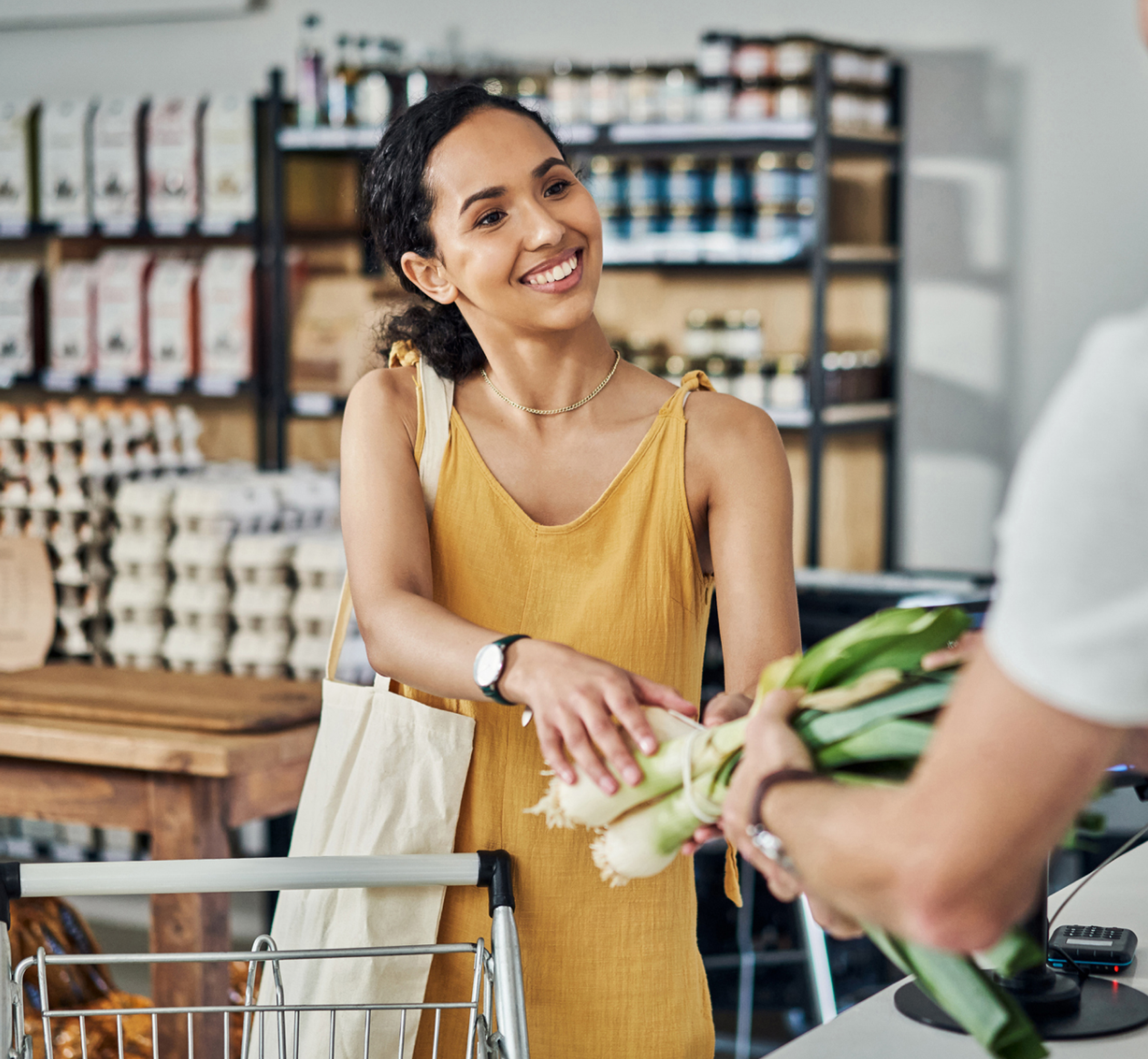 Smiling woman in a yellow dress accepts a bunch of fresh leeks from a cashier in a grocery store.