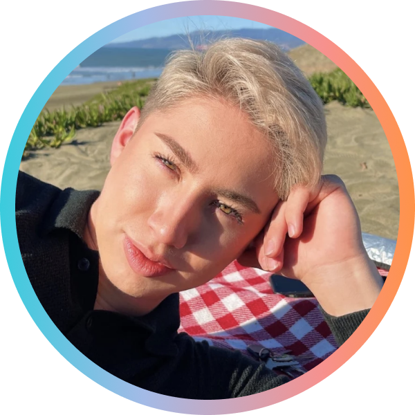 A person with light blonde hair rests their head on their hand, smiling while sitting on a beach with a picnic blanket. The background features sand and greenery with a view of the ocean.