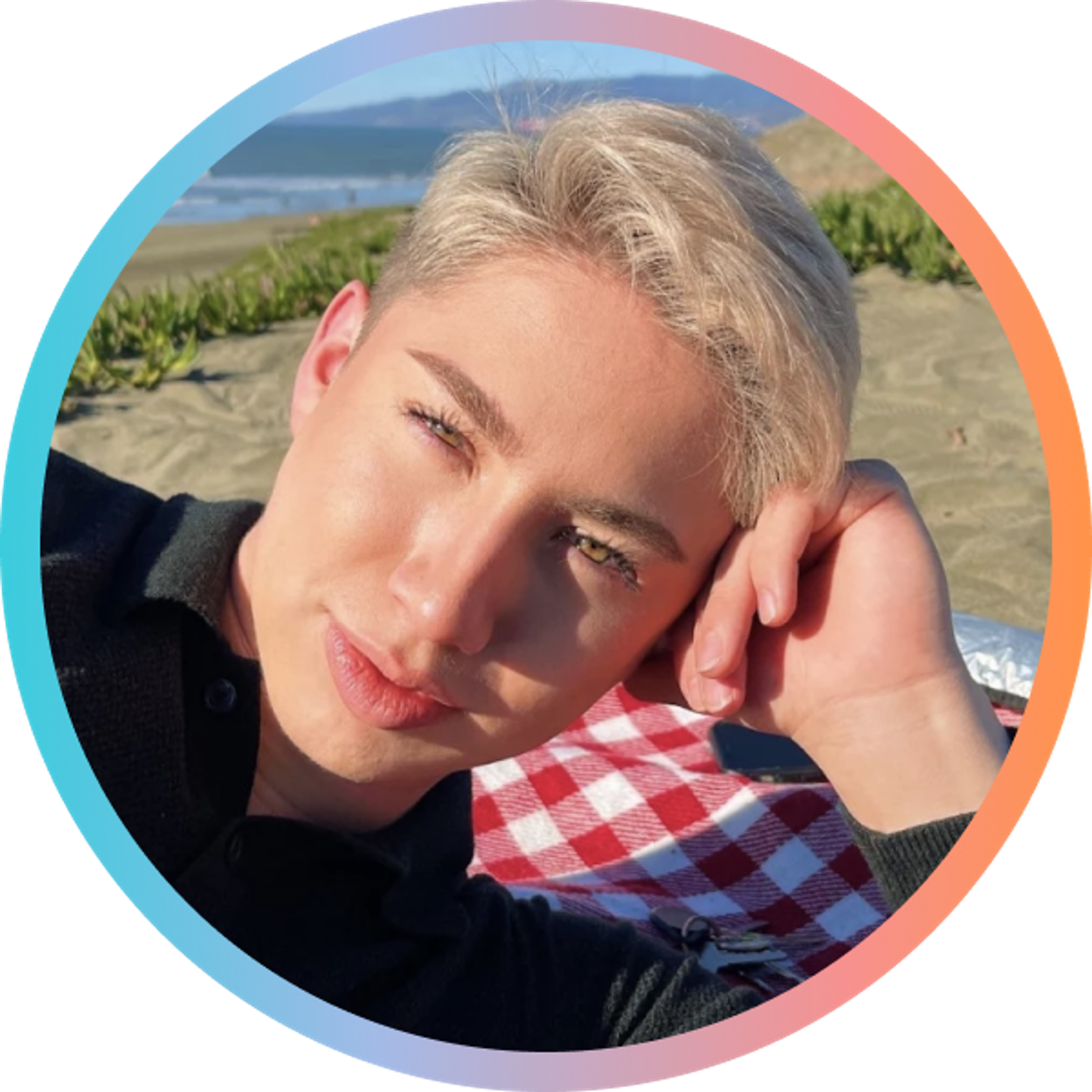 A person with light blonde hair rests their head on their hand, smiling while sitting on a beach with a picnic blanket. The background features sand and greenery with a view of the ocean.