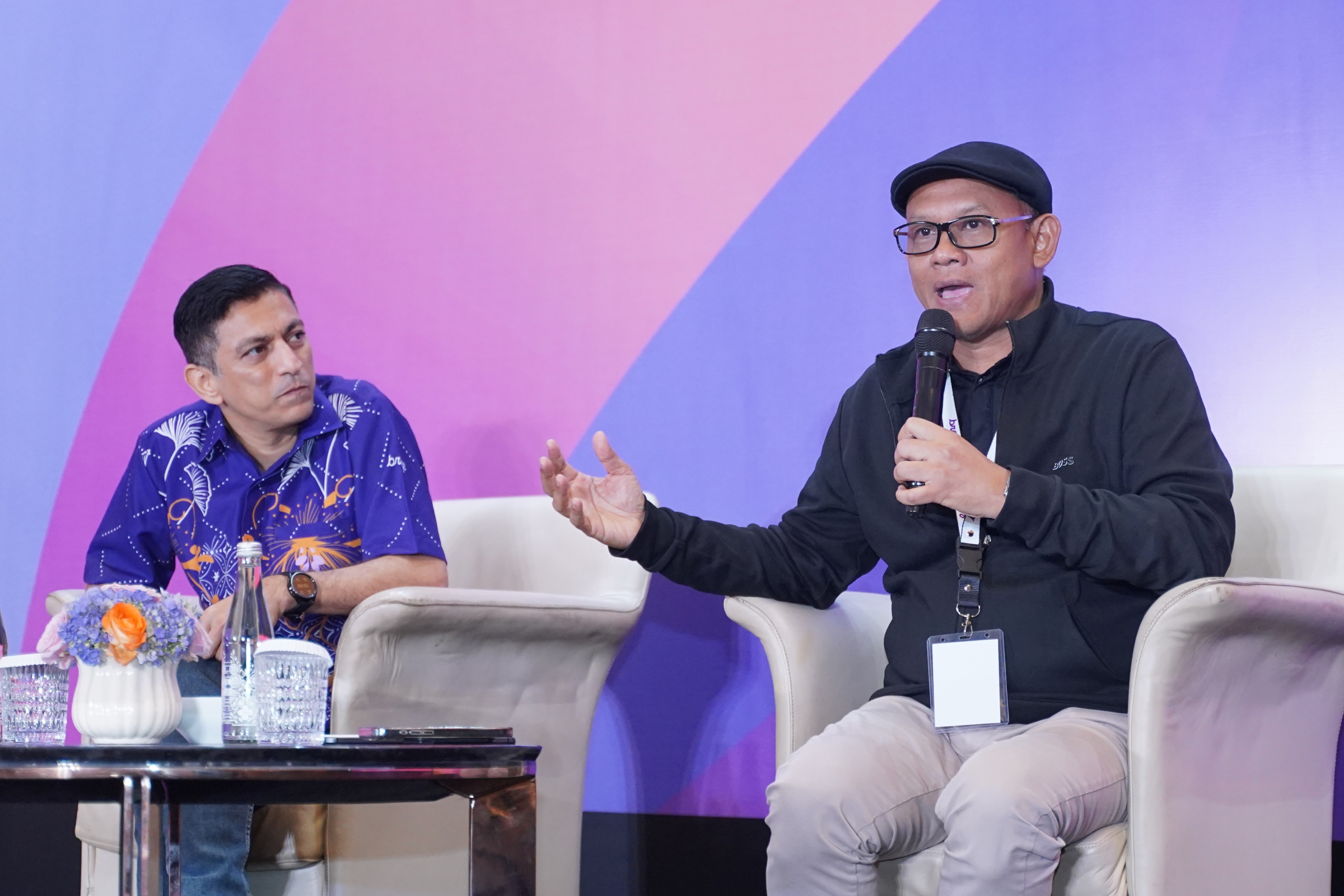A man in a black cap speaks into a microphone and gestures during a panel discussion as another seated man listens.