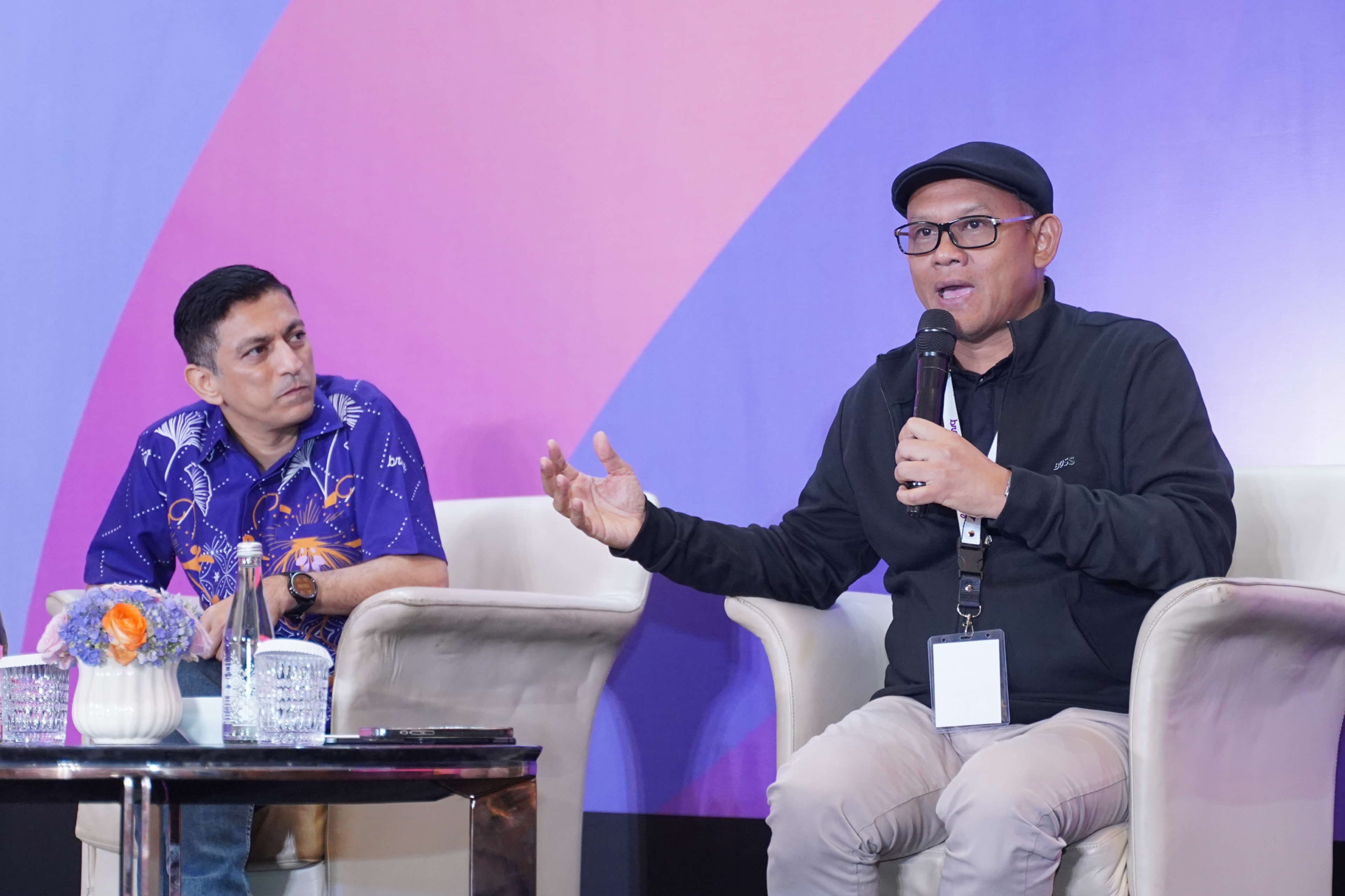 A man in a black cap speaks into a microphone and gestures during a panel discussion as another seated man listens.