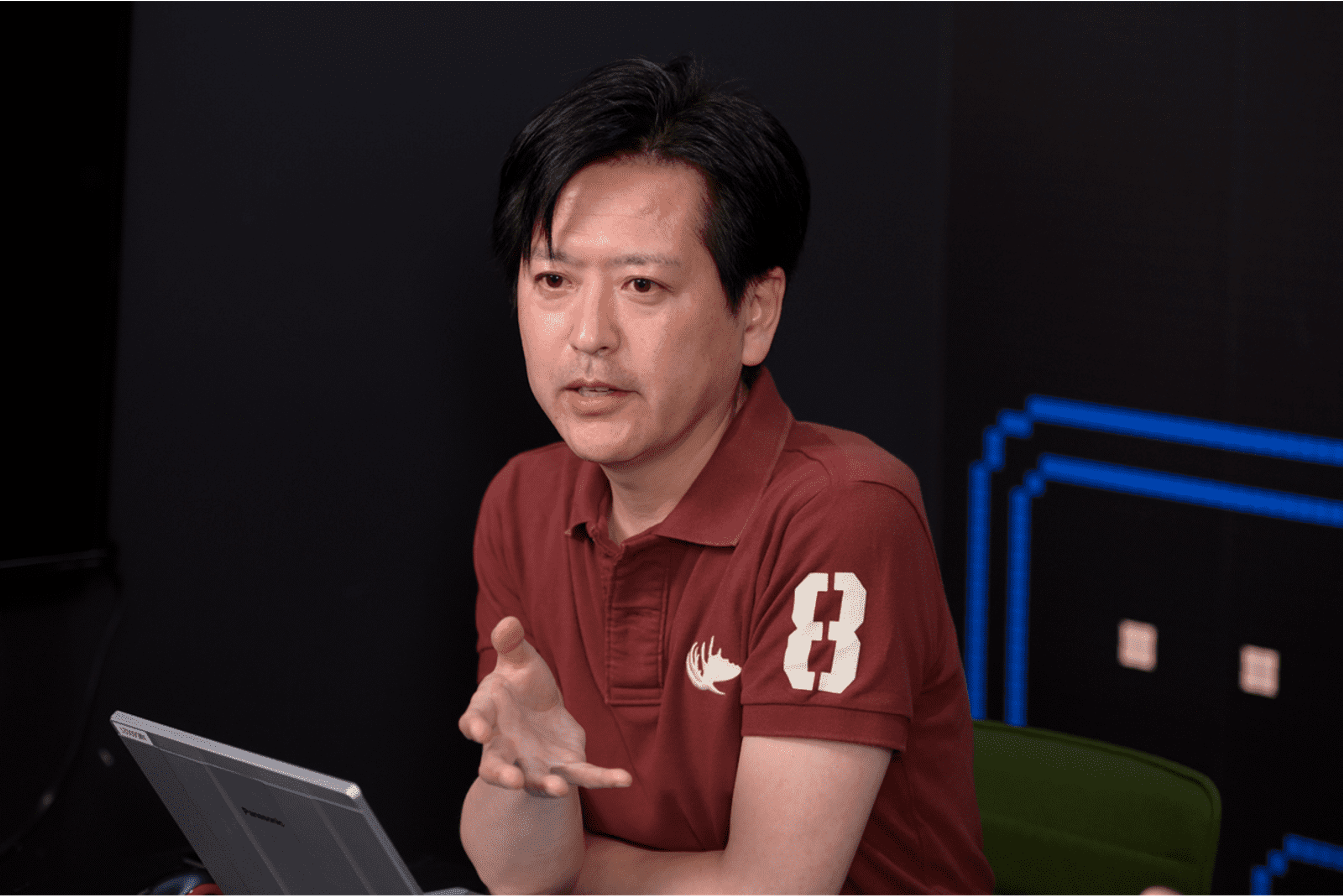 バンダイナムコ社員の一人が、ゲーム画面の背景の前で腕をくんで座ってインタビューに答えている様子