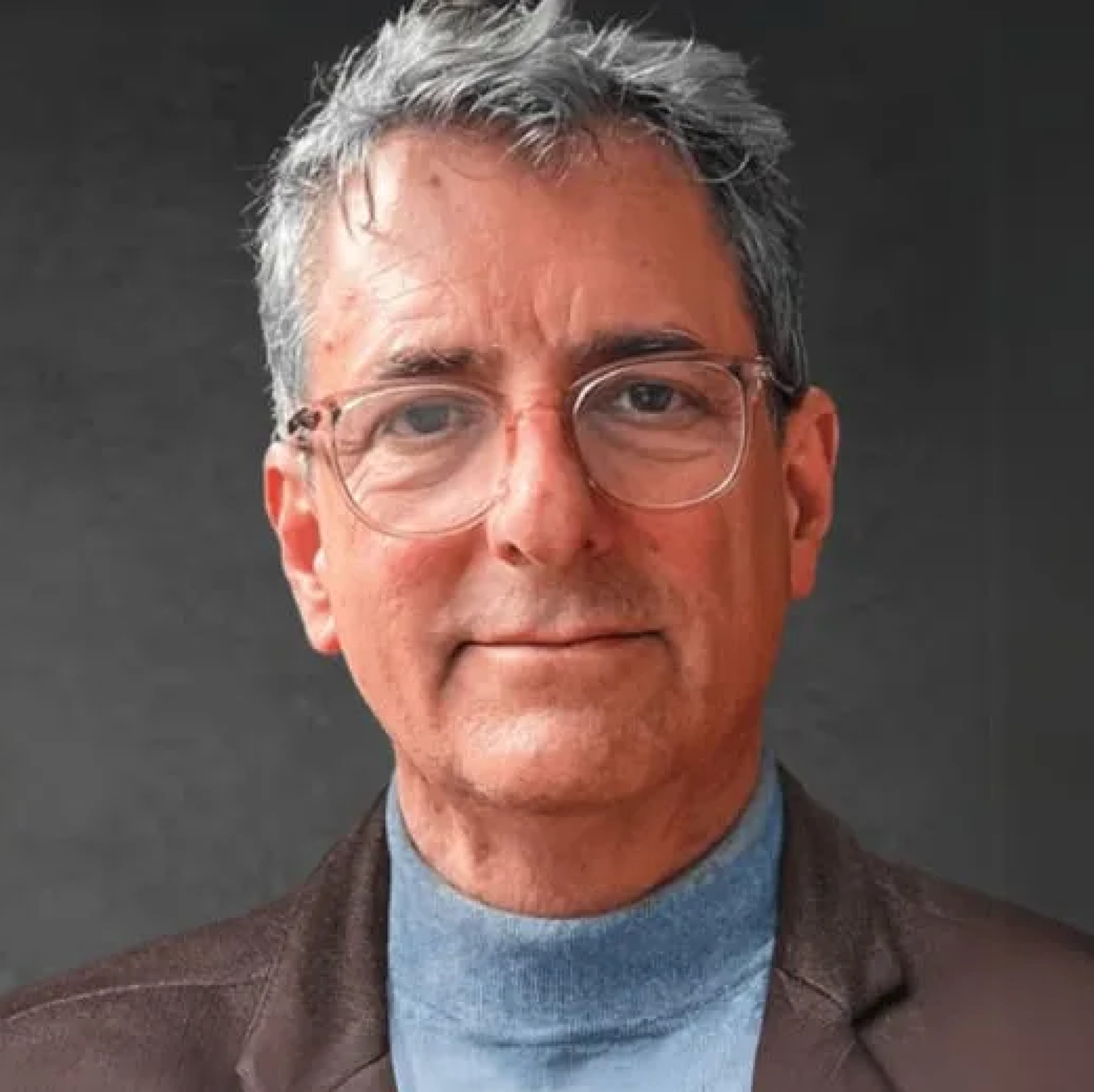 A middle-aged man with gray hair and glasses smiles at the camera, wearing a brown blazer over a blue turtleneck. The background is dark and neutral.