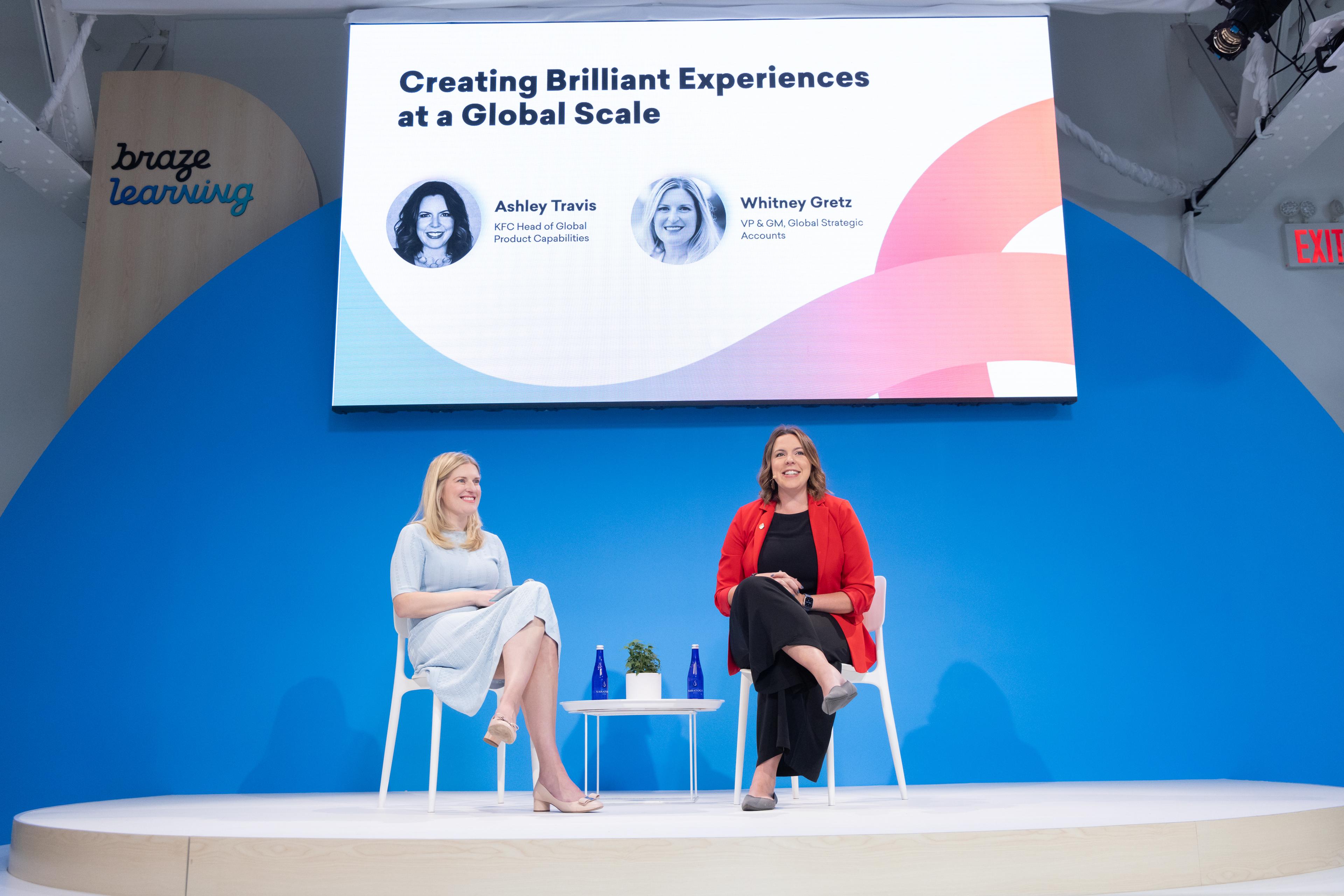 Two women sit on stage in front of a large screen displaying the title "Creating Brilliant Experiences at a Global Scale." The setting features a blue backdrop and a modern design.