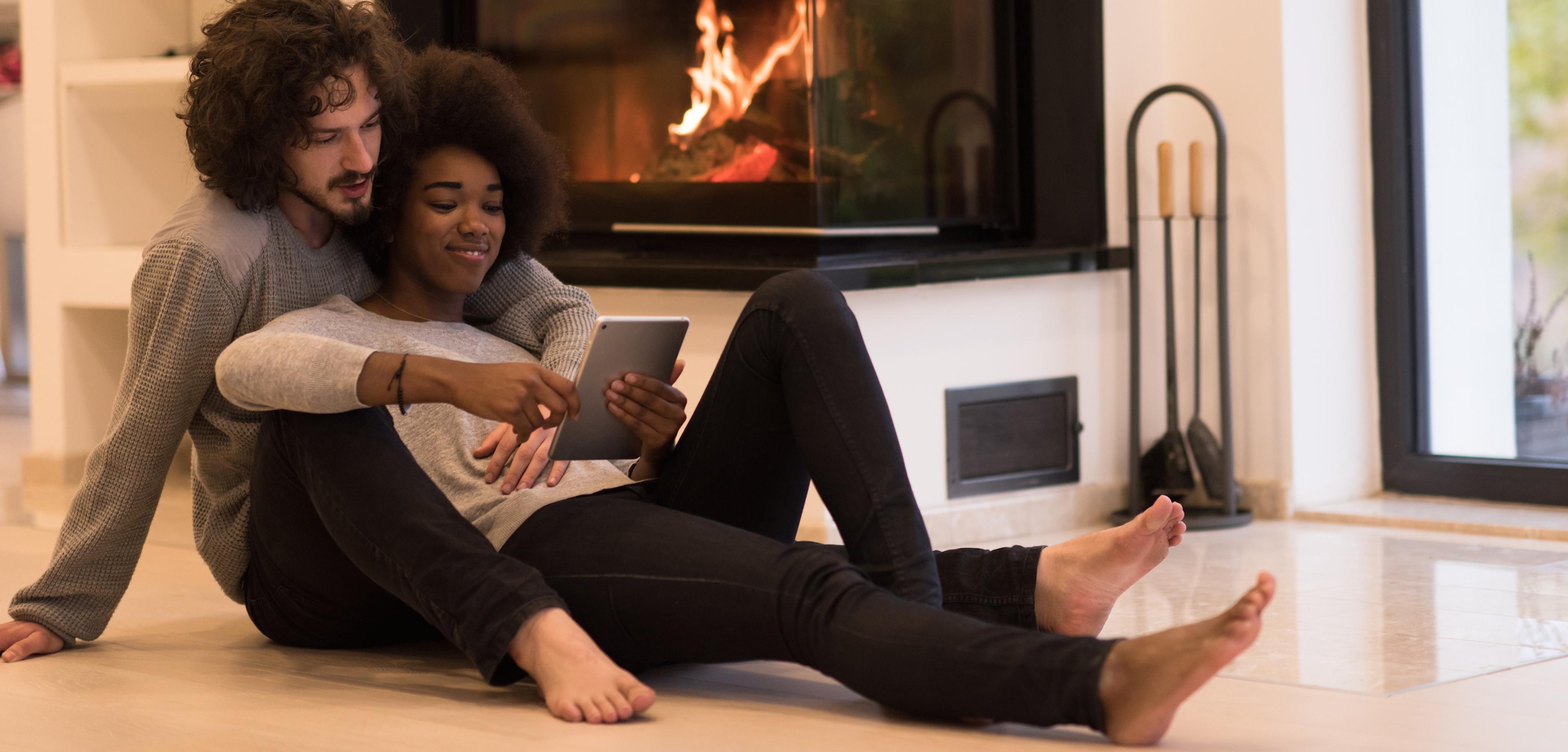 A couple sits on the floor by a fireplace, sharing a tablet. They appear relaxed and engaged with each other.