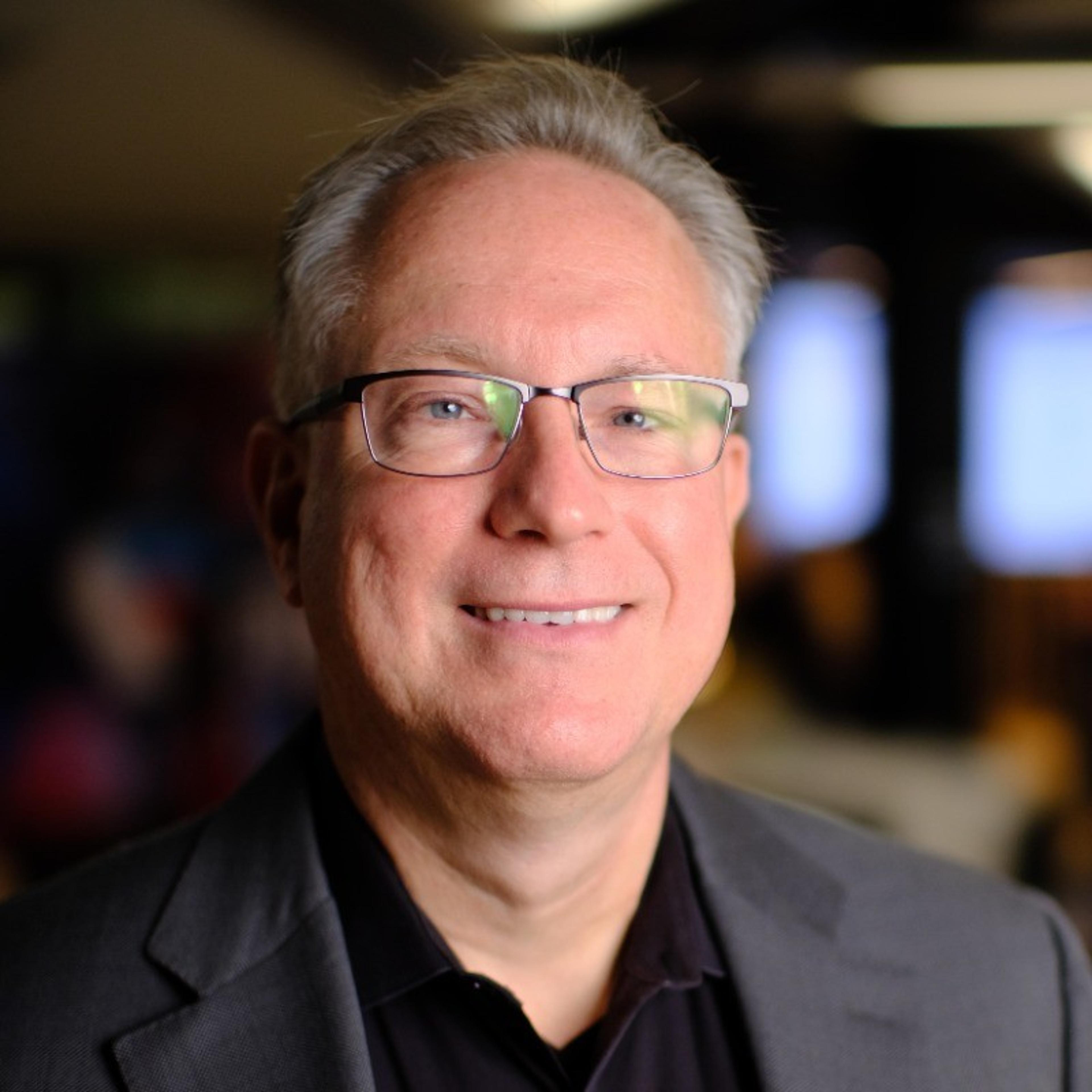 A smiling man with glasses and gray hair, wearing a dark blazer, stands in a well-lit indoor setting.