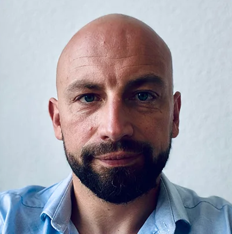 A man with a bald head and a beard is looking directly at the camera, wearing a light blue shirt. The background is plain and light-colored.