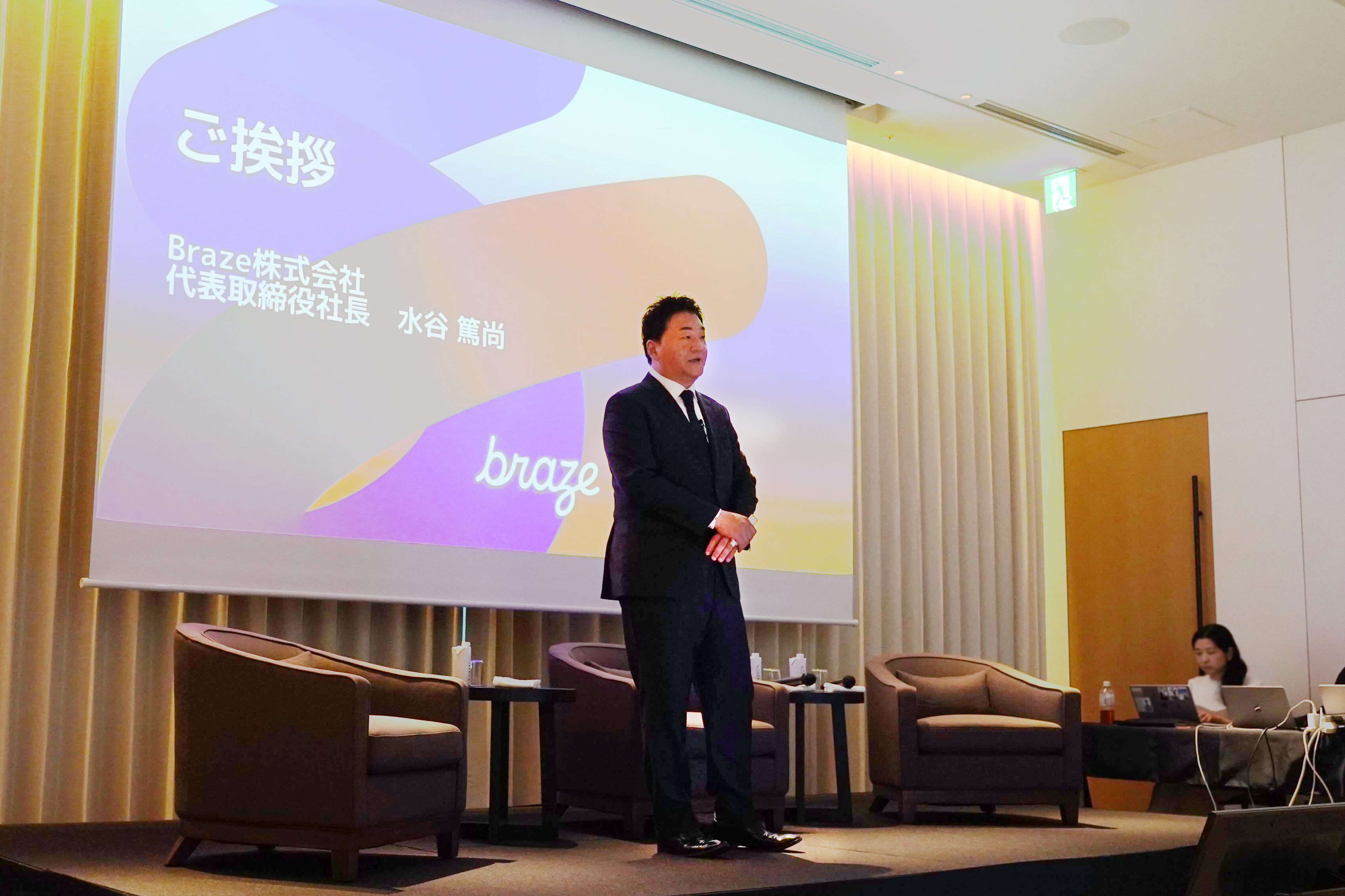 ブラゼ株式会社の代表取締役社長がプレゼンテーションを行っている様子。背景には会社のロゴとスライドが表示されている。