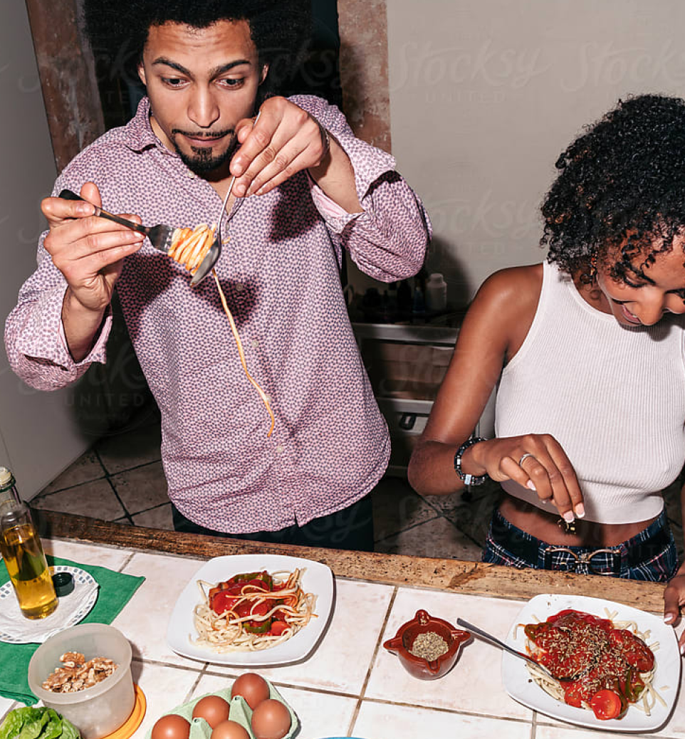 Um homem serve espaguete em um prato enquanto uma mulher ao lado adiciona tempero em sua refeição. Ambos estão em uma cozinha.