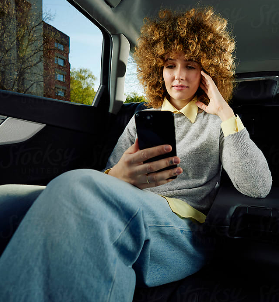 A young woman with curly hair sits in the backseat of a car, looking at her smartphone with a thoughtful expression. She wears a gray sweater and light blue pants.