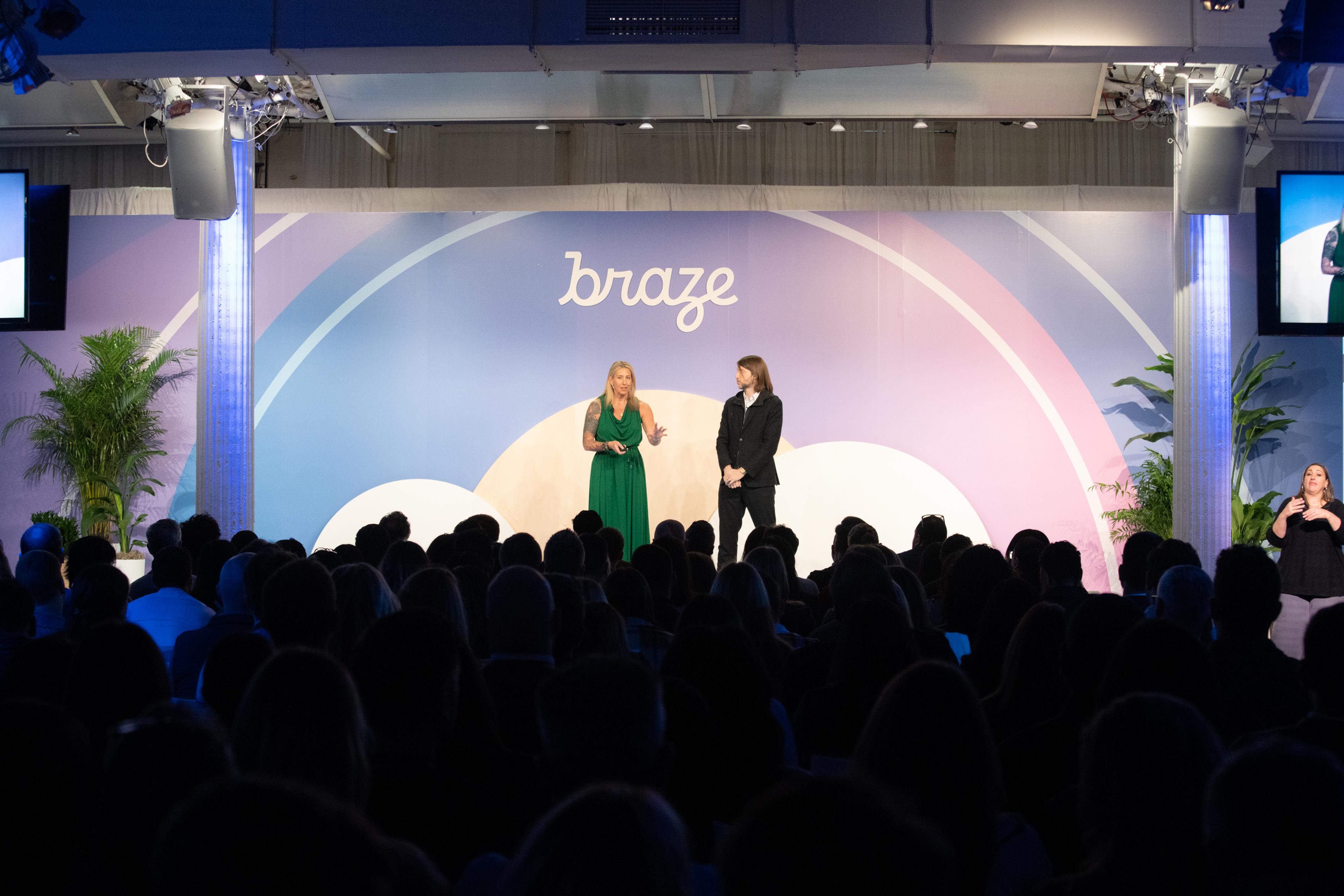 A speaker in a green dress stands on stage next to another speaker in a black suit, addressing an audience at a Braze event. The backdrop features the Braze logo and colorful design elements.