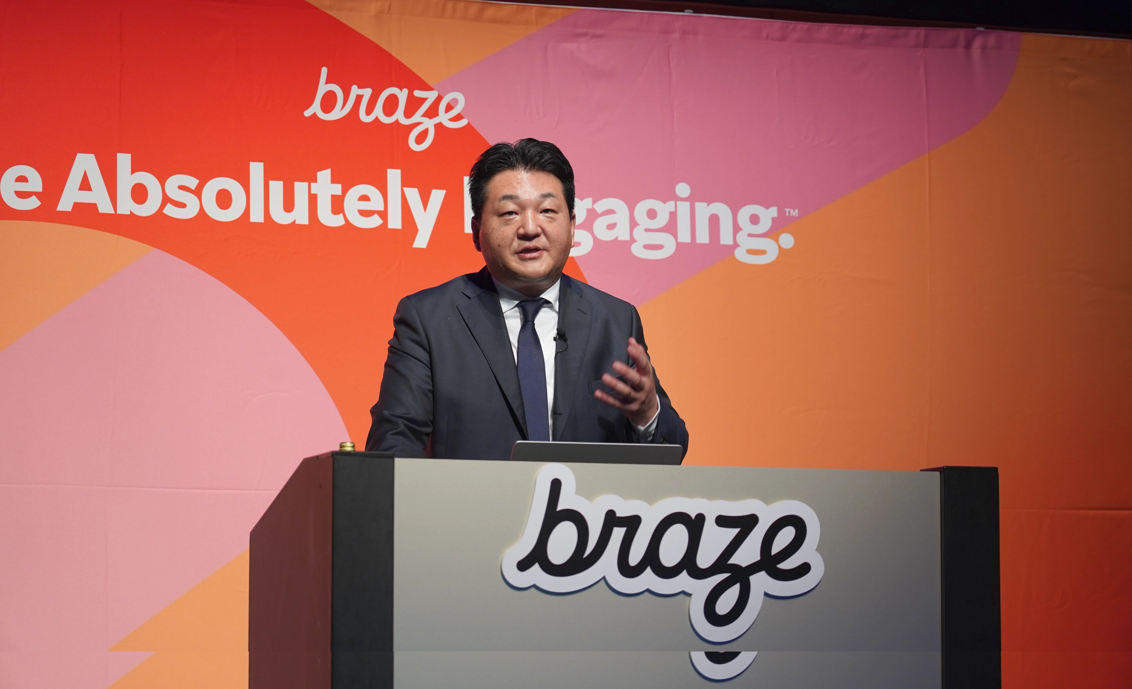 男性が「braze」のロゴの前でプレゼンテーションを行っています。背景にはカラフルなデザインがあります。