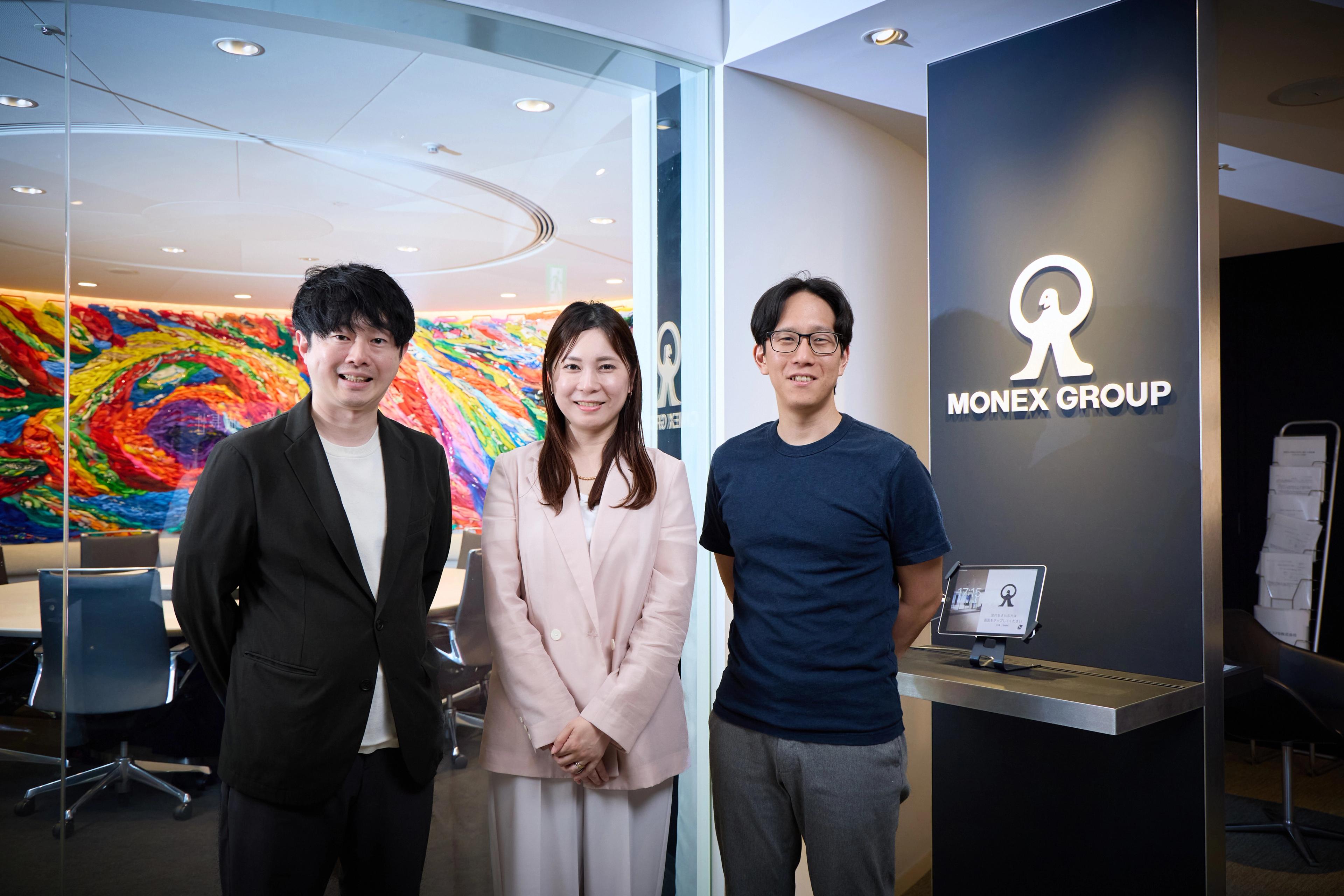 インタビューを受けたmonex groupsの社員3人がロゴの前に立っている