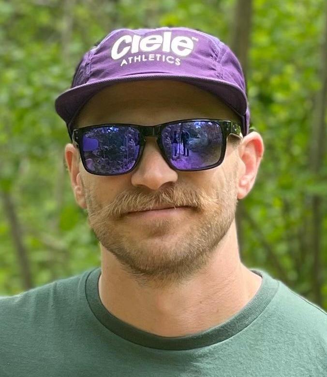 A man with a mustache wears sunglasses and a purple cap with "Ciele Athletics" printed on it, standing in a green, wooded area.