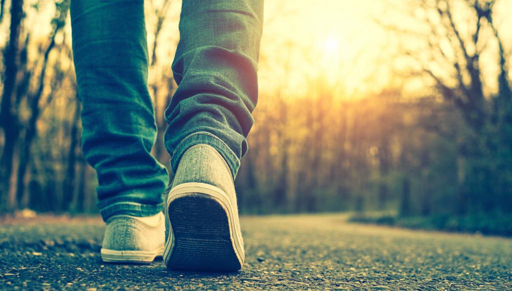 A person walks down a tree-lined path at sunset, wearing blue jeans and white sneakers. The warm light creates a serene atmosphere.