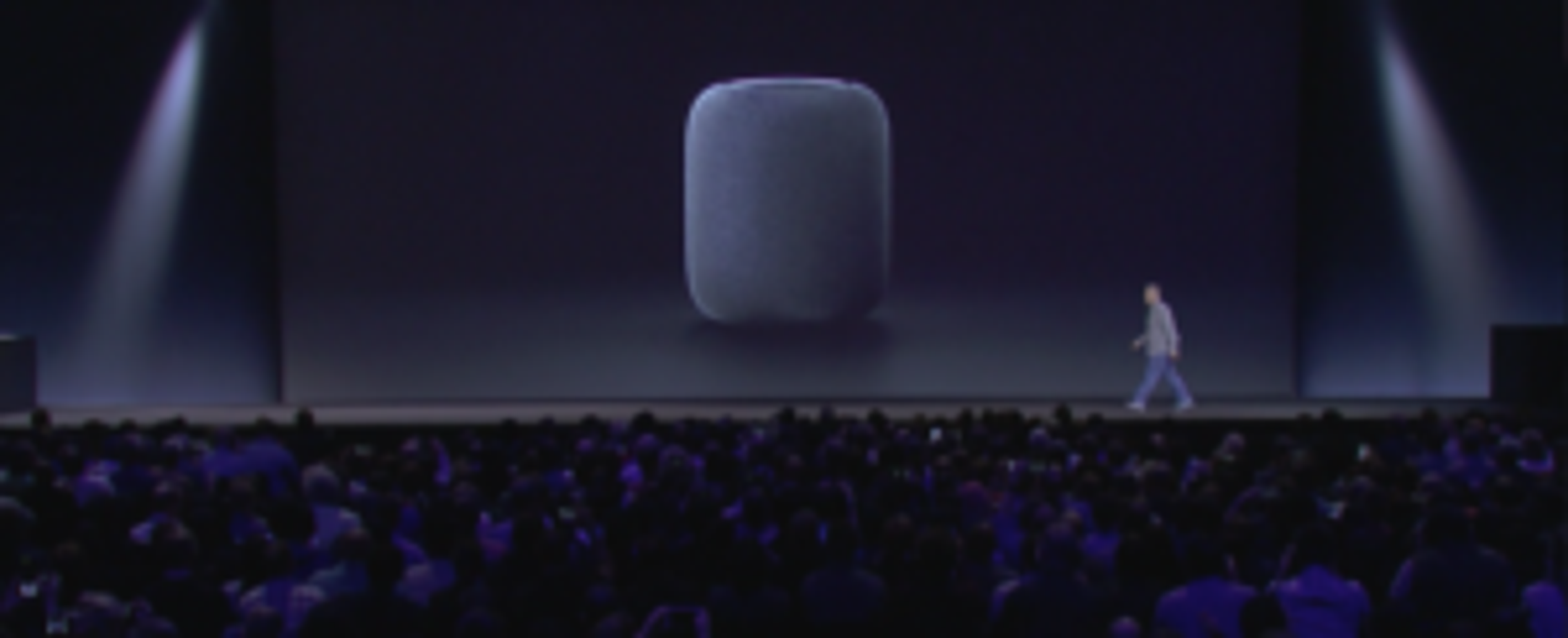 A speaker device is displayed on a large screen while a presenter walks across the stage in front of an audience. The setting is dimly lit with a focus on the speaker.