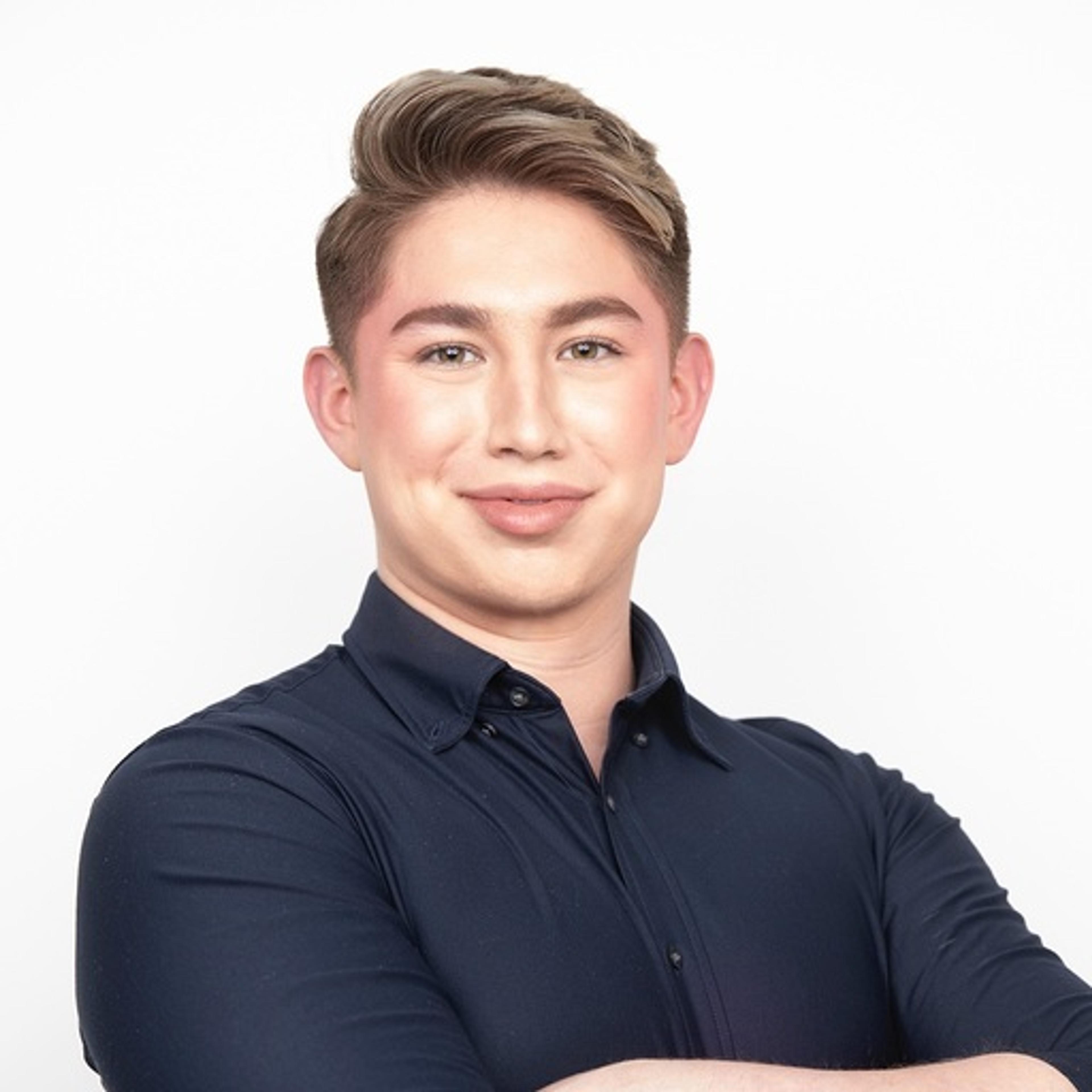 A young person with light brown hair and makeup smiles confidently while crossing their arms, wearing a dark blue shirt against a white background.