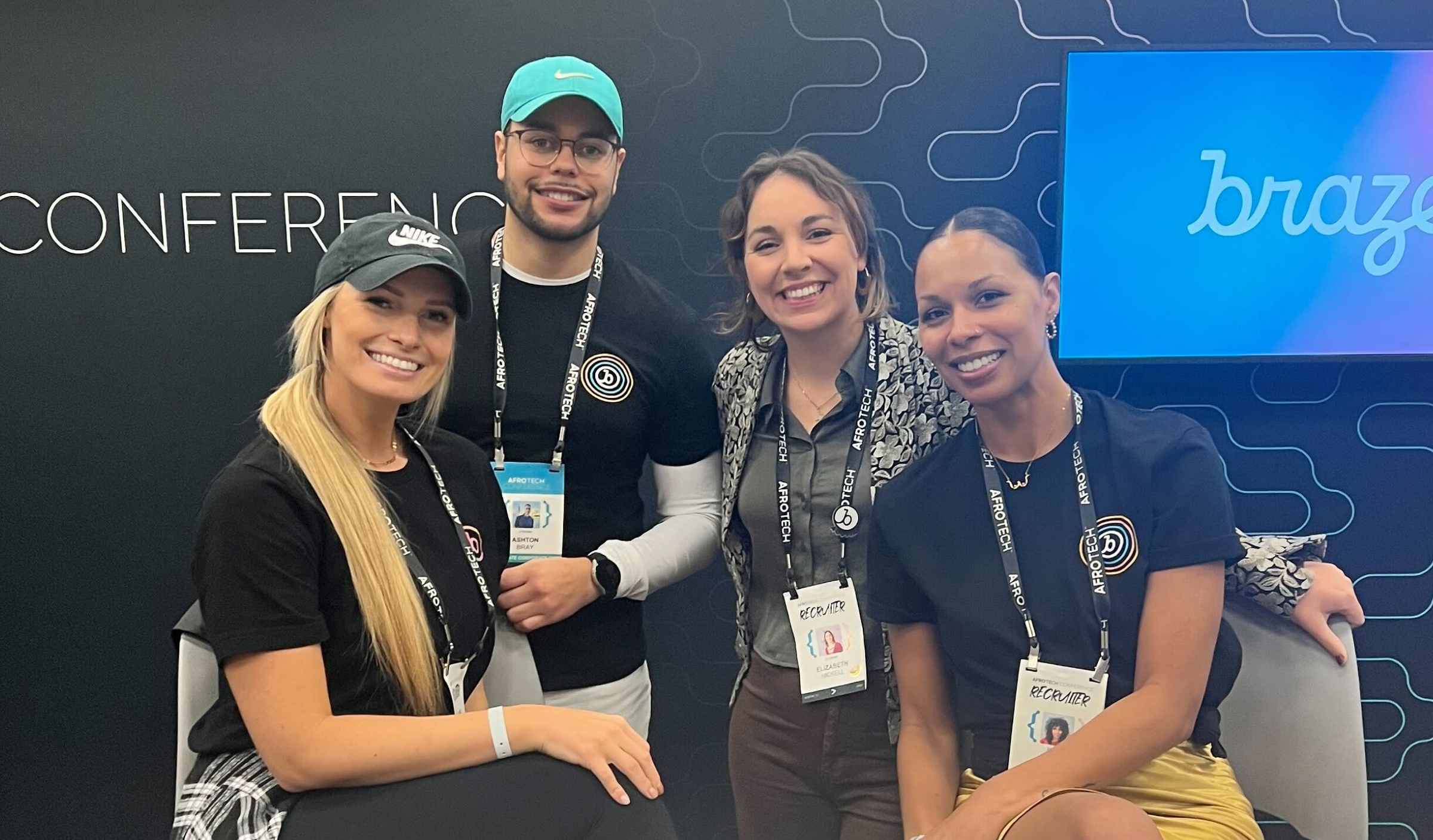 Four people pose together at a conference booth, smiling and wearing branded attire. The background features a logo and a digital display.