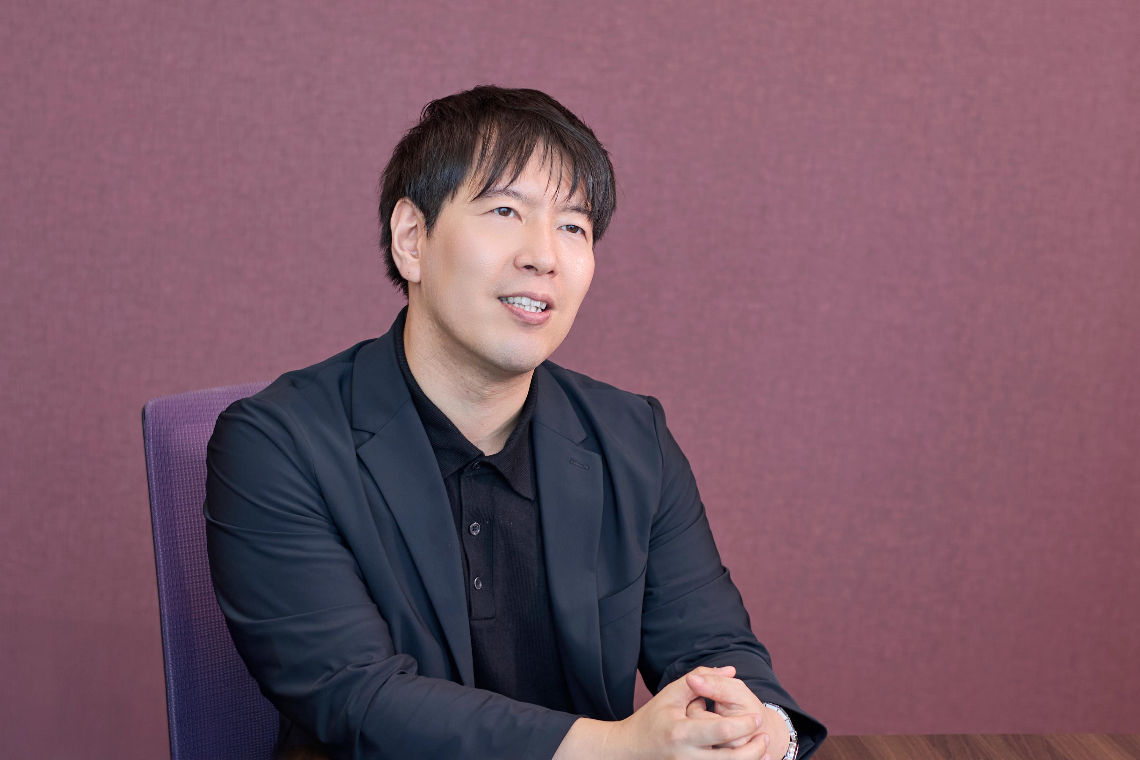 a man in a suit and black shirt is sitting at a table with his hands folded .