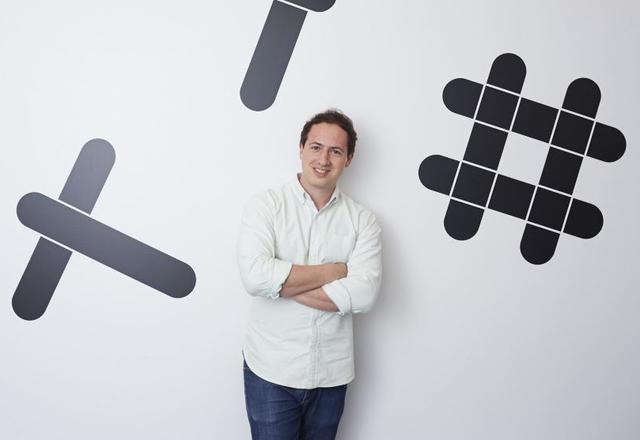 A person stands with arms crossed in front of a wall featuring large black graphic symbols. They are smiling and wearing a light-colored shirt.